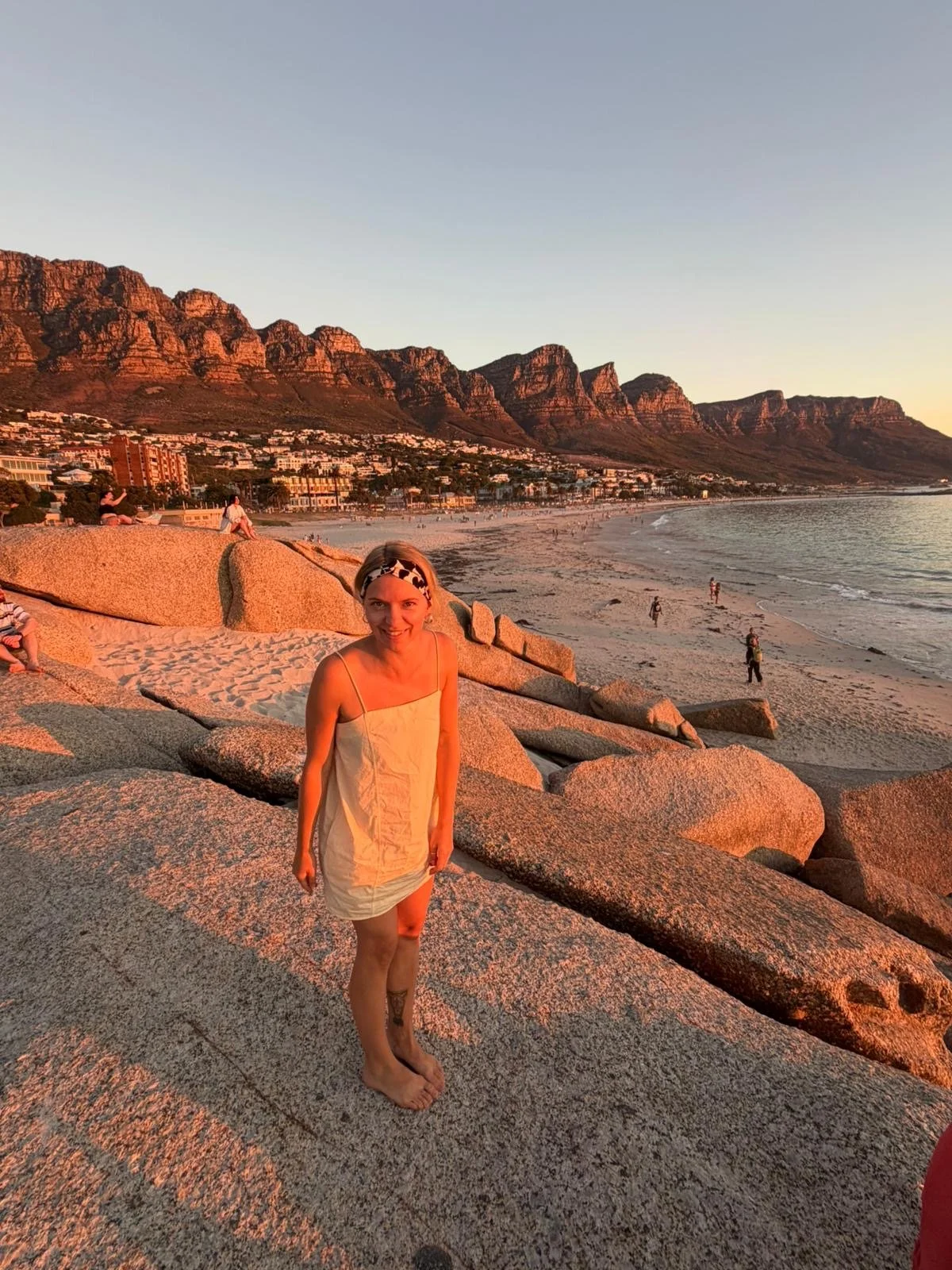 Eine Frau in einem weißen Kleid und Kopfbedeckung steht barfuß auf großen Granitfelsen am Strand während des Sonnenuntergangs, im Hintergrund die Berge und das Meer.