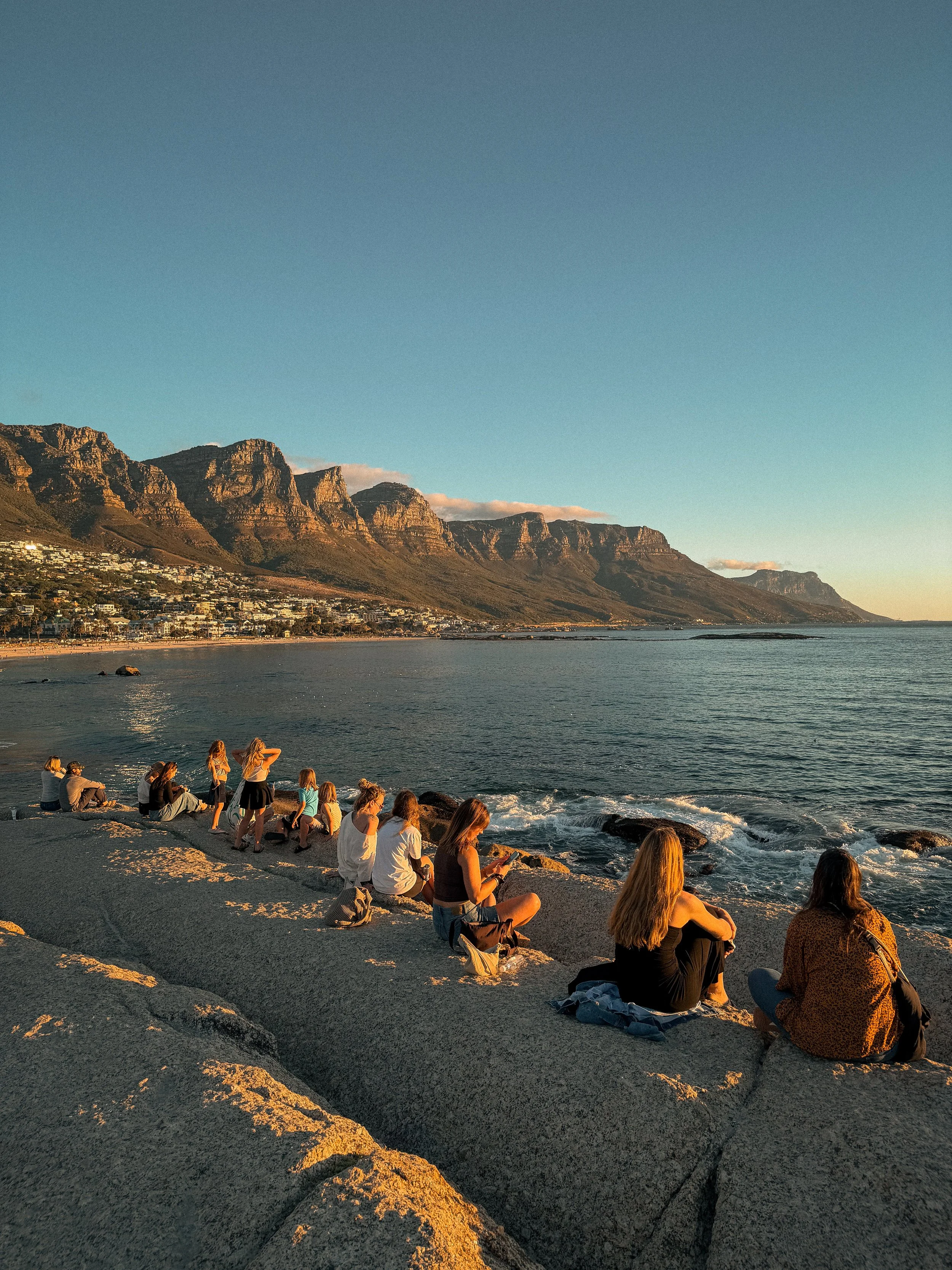 Menschen sitzen am Strand während des Sonnenuntergangs mit Blick auf die an der Küste gelegenen Stadt und die Berge im Hintergrund.