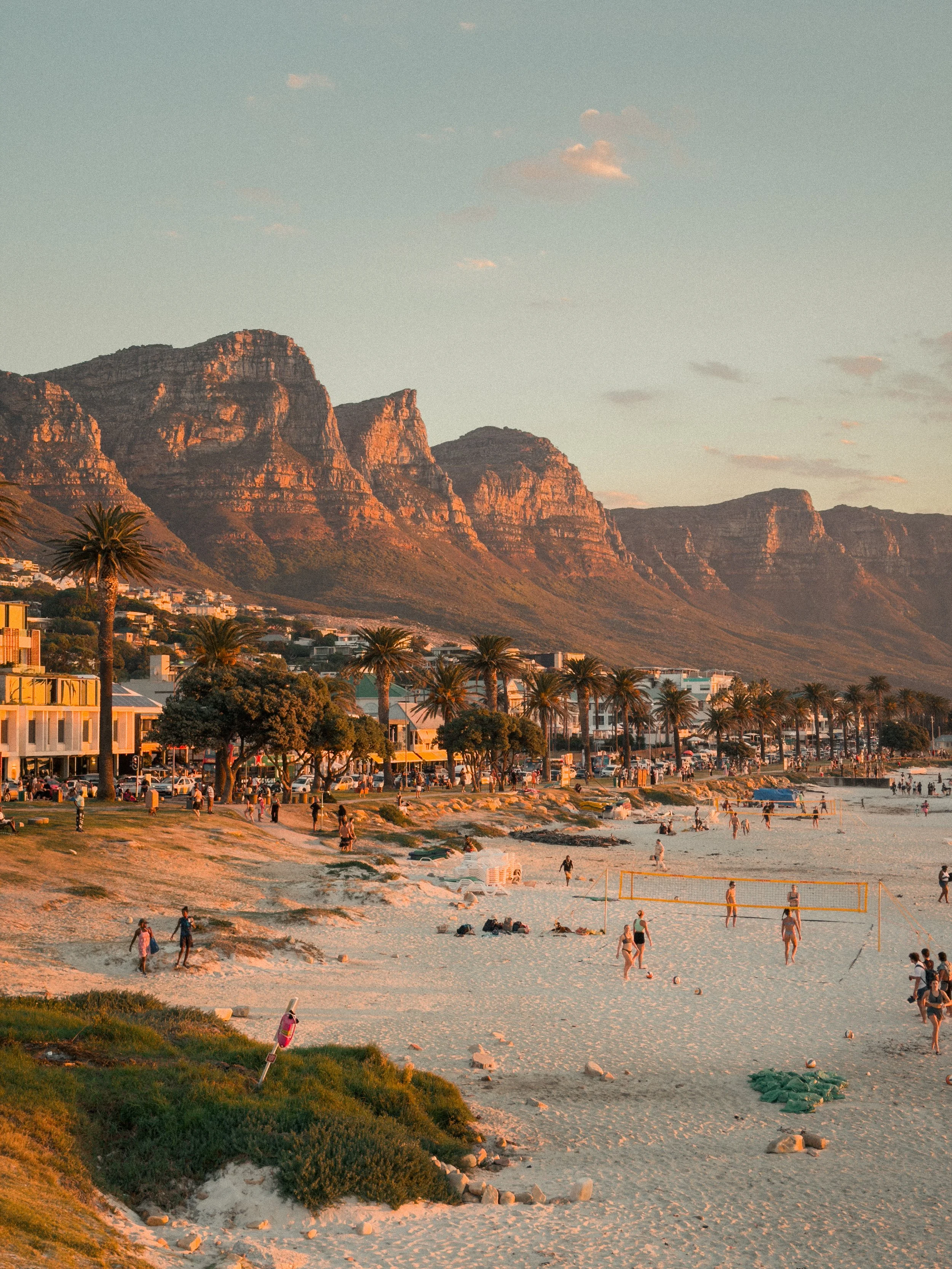 Strand bei Sonnenuntergang mit Menschen, Palmen, Gebäuden im Vordergrund und Bergen im Hintergrund.