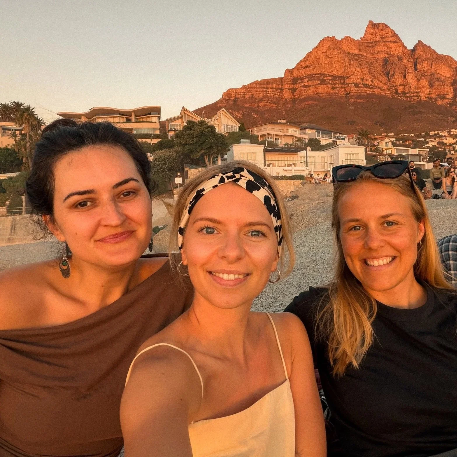 Drei Frauen posieren für ein Selfie am Strand vor einer Bergkulisse bei Sonnenuntergang.