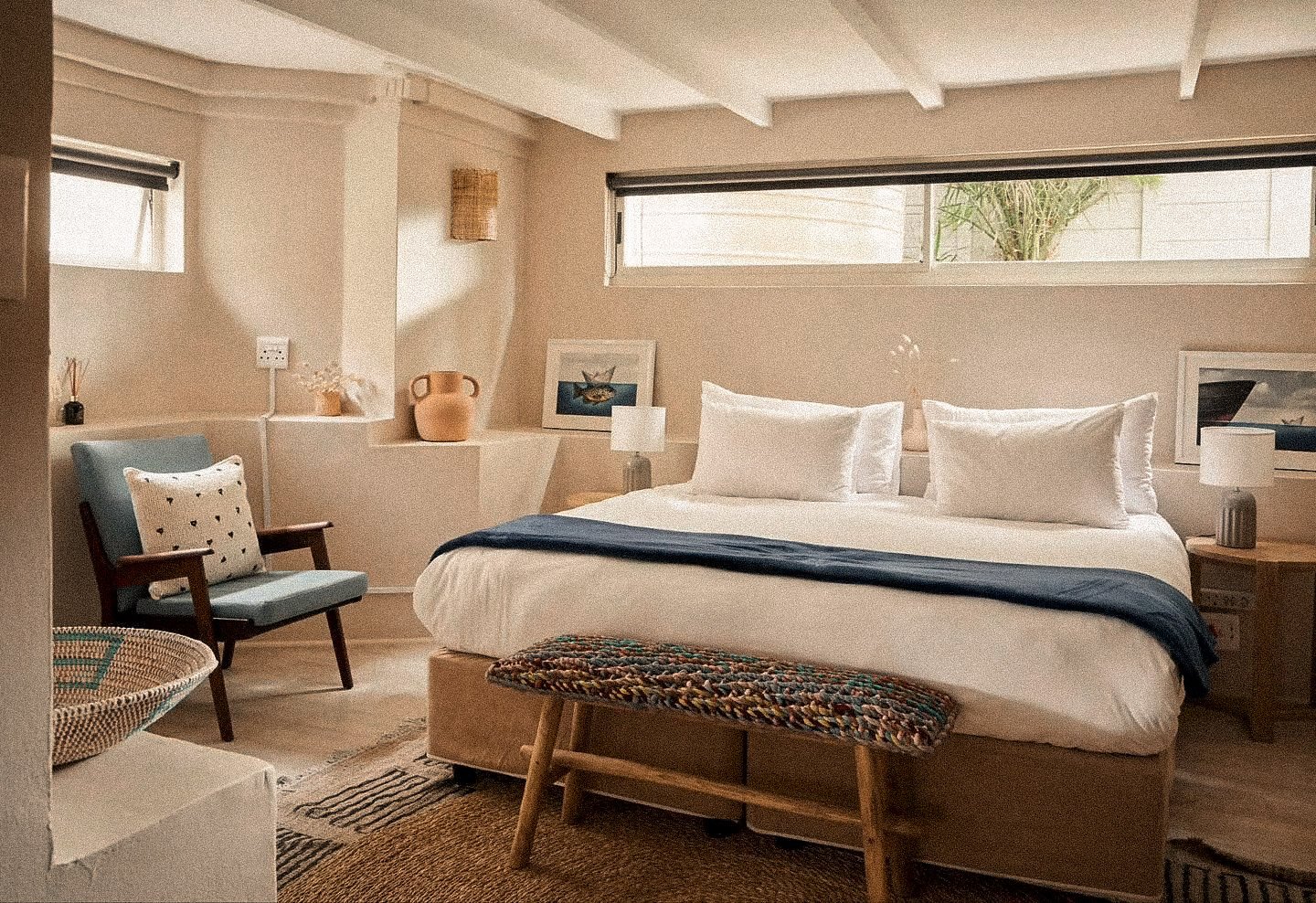 Ein gemütliches Schlafzimmer mit einem großen weißen Bett, einer blauen Decke, einem bunten Hocker am Fußende, einem blauen Armlehnenstuhl mit Kissen, seitlichen Nachttischen mit Lampen, einem Fenster mit Blick nach draußen, und verschiedenen Dekorationsgegenständen.