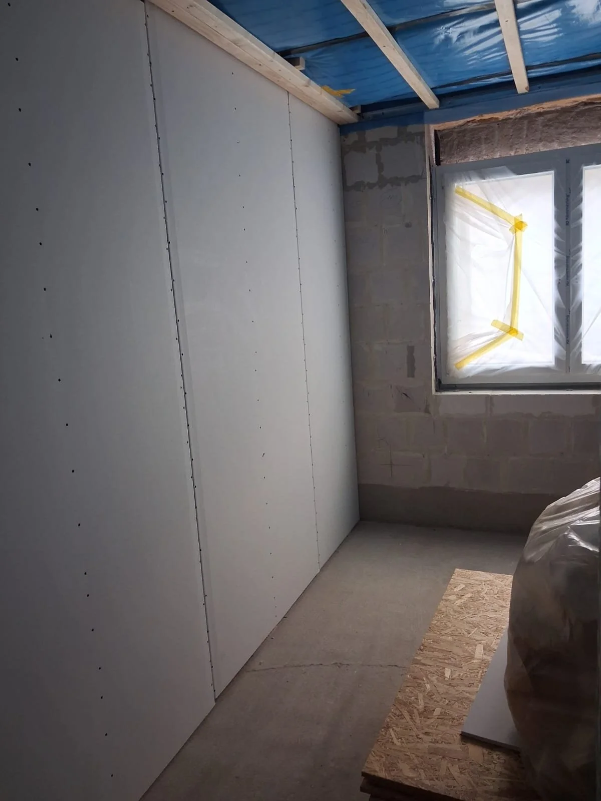 Innenansicht eines unfertigen Raumes mit zweiflügeliger Türe auf der linken Seite, eine Wand mit Gipsplatten, eine Fensteröffnung mit Schutzfolie und gelber Malerkreppband, unbedeckte Holzdecke, unvollständiger Putz auf den Wänden, auf dem Boden befi