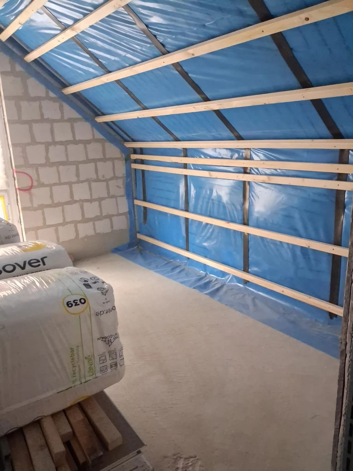 Innenraum eines unvollendeten Raumes mit Isolierung und Holzrahmen an der Decke, Baustaub und Baumaterialien im Vordergrund.