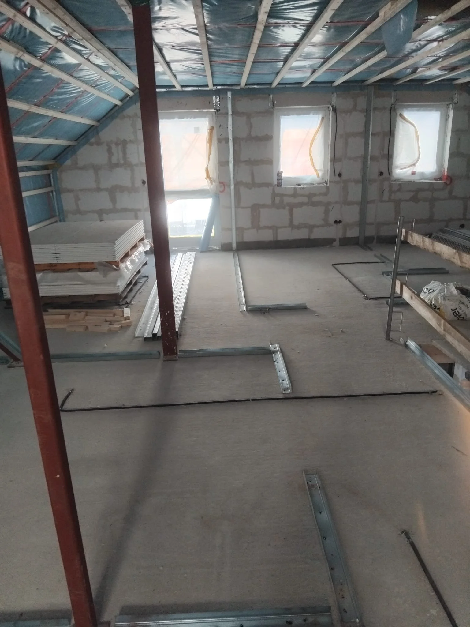 Innenansicht eines Rohbaus, Wände mit Ziegelsteinen, mehrere Fenster, im Raum sind Baumaterialien und Metallrahmen für eine Decke oder einen Boden