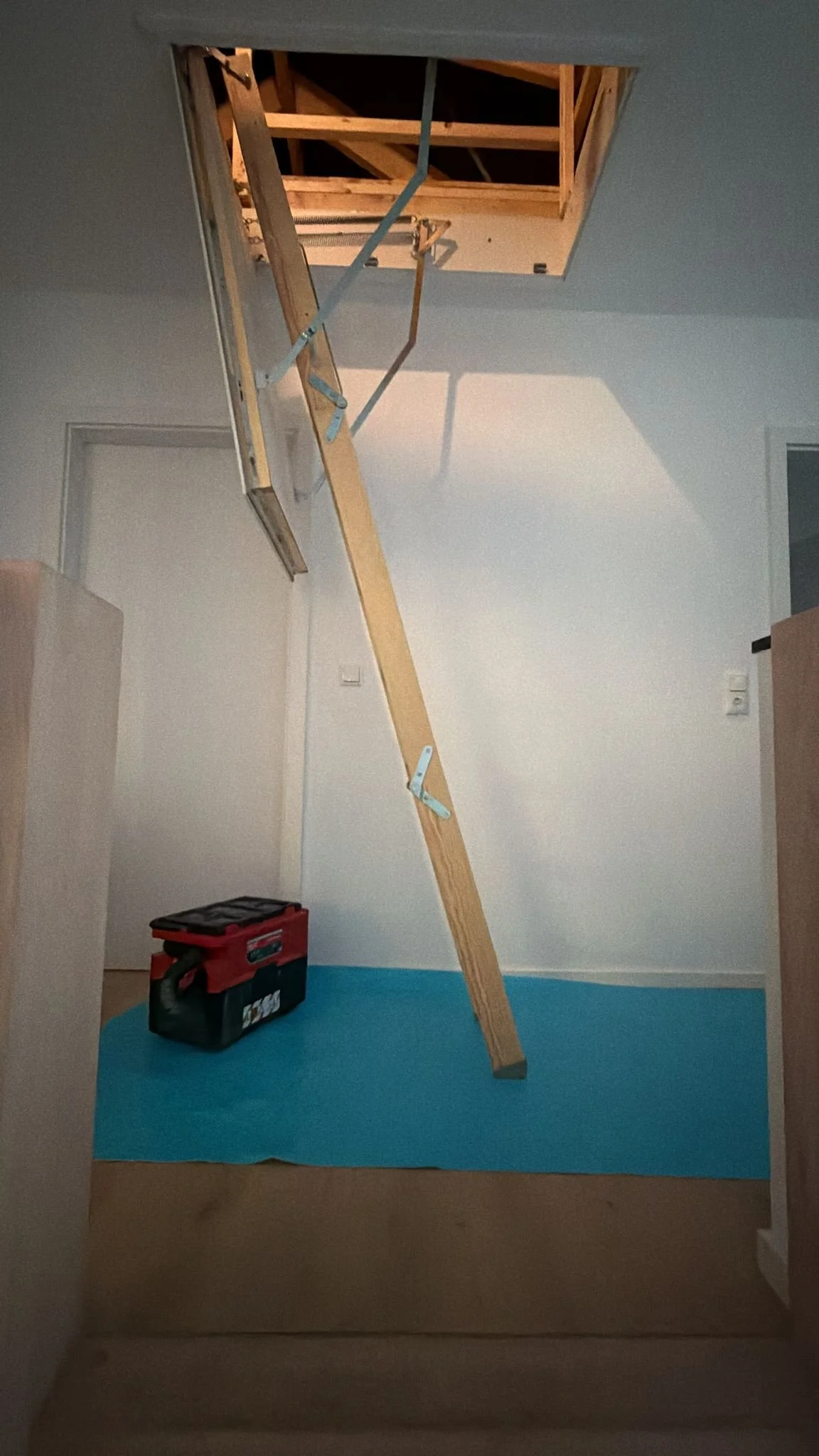 Bauarbeiten im Innenraum, Holztreppe führt zu einem Dachboden, blauer Bodenbelag, Werkzeugkoffer auf dem Boden.