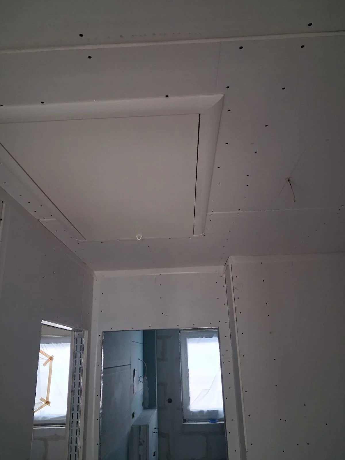 Innenraum eines im Bau befindlichen Raumes mit Trockenbauwänden und einer schlecht beleuchteten Decke, das Fenster ist mit weißem Schutzmaterial bedeckt.