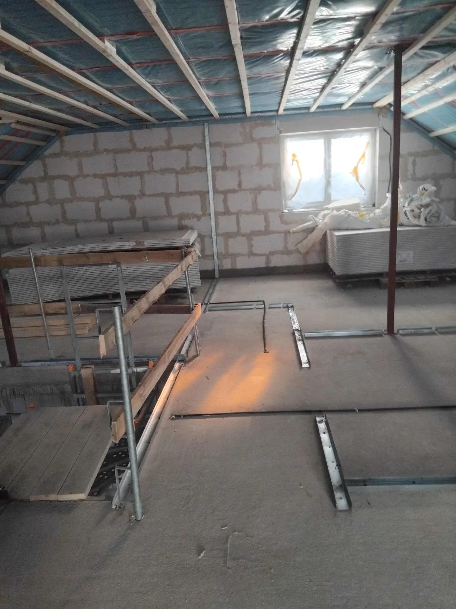 Innenraum eines unvollständigen Dachgeschosses mit sichtbaren Dachbalken, ungeputzter Ziegelmauer, einem Fenster mit Plastikfolie und Heizkörper darunter, sowie Baugerüsten und Baumaterialien.