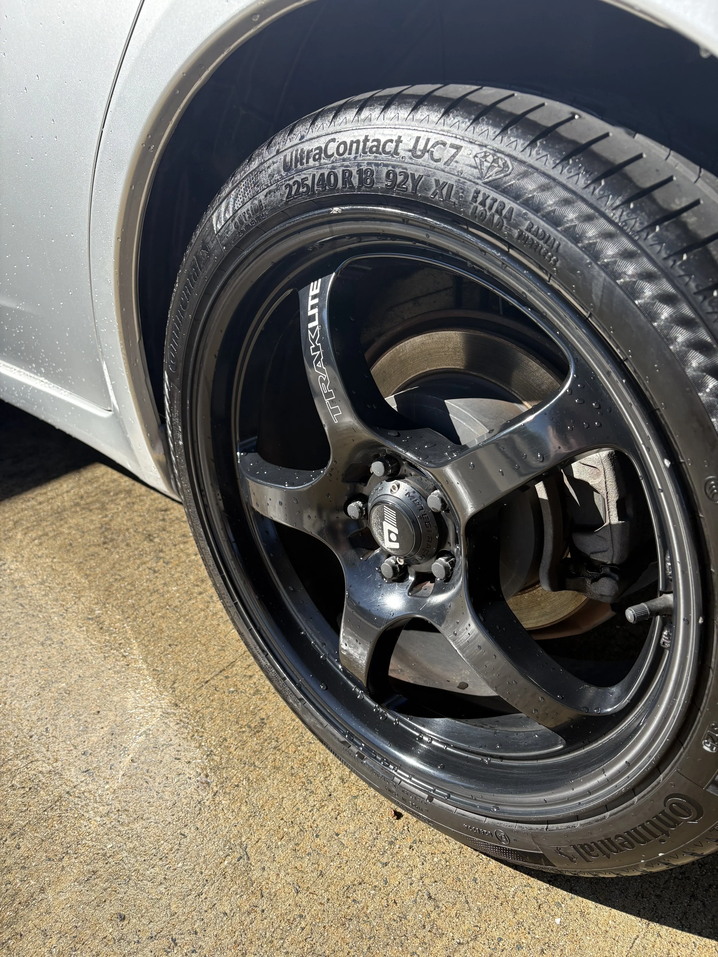 Close-up of a car tire and rim, showing the tire's detailed sidewall with markings and a partial view of the car's silver wheel well on a concrete surface.