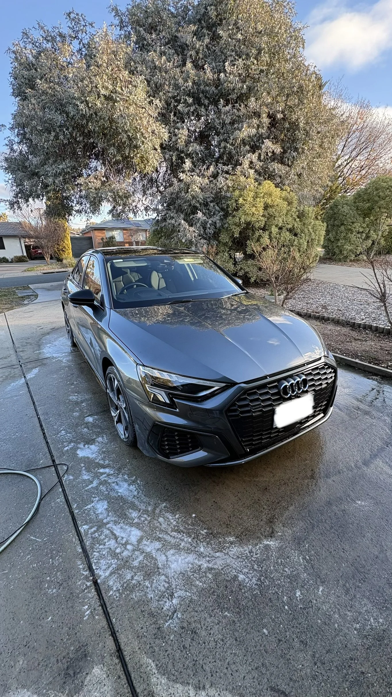 Gray Audi car parked on a wet driveway with soap suds, trees, and a suburban neighborhood in the background.