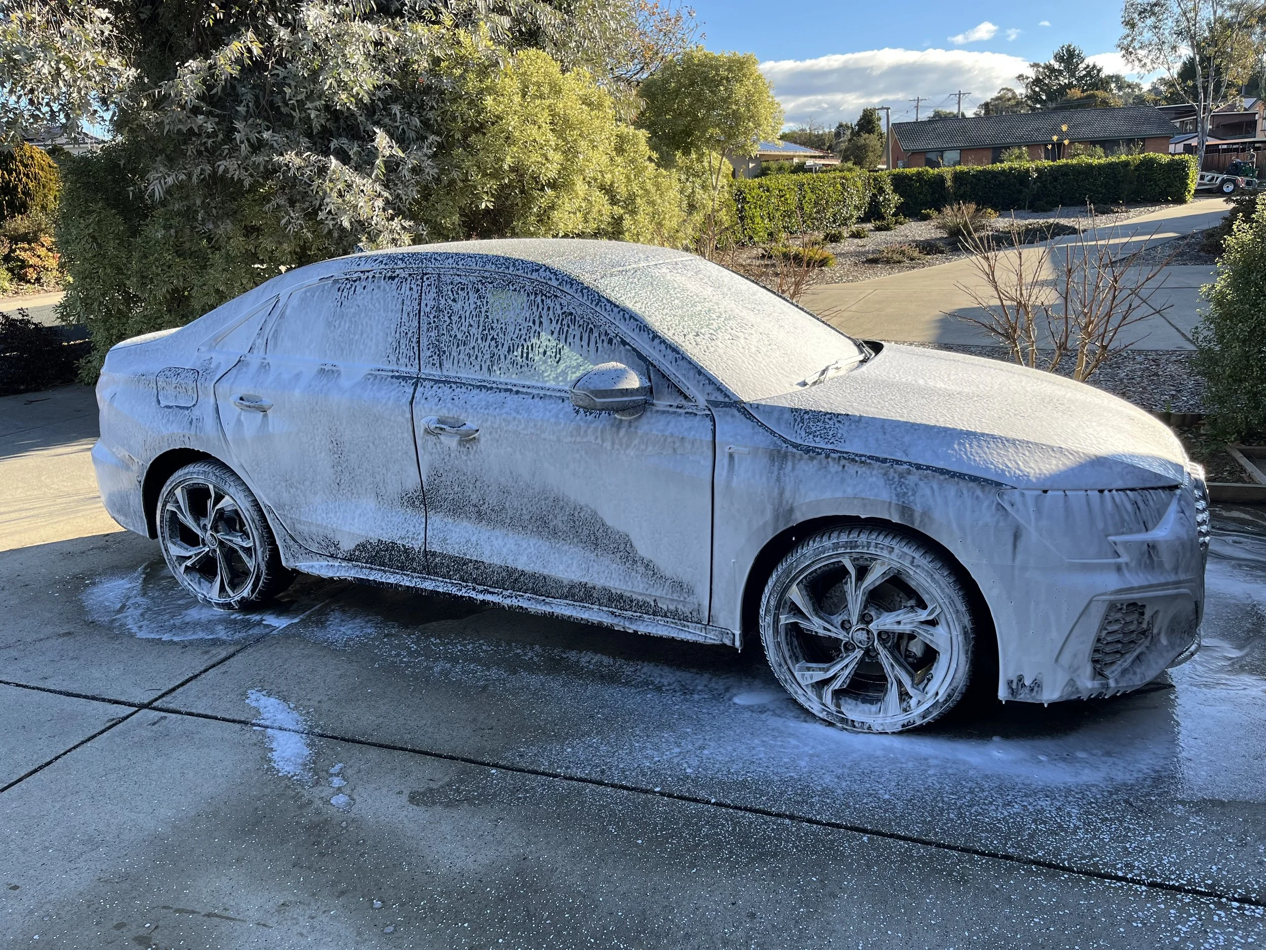 A white car covered in soap suds during a car wash on a driveway with trees and houses in the background.