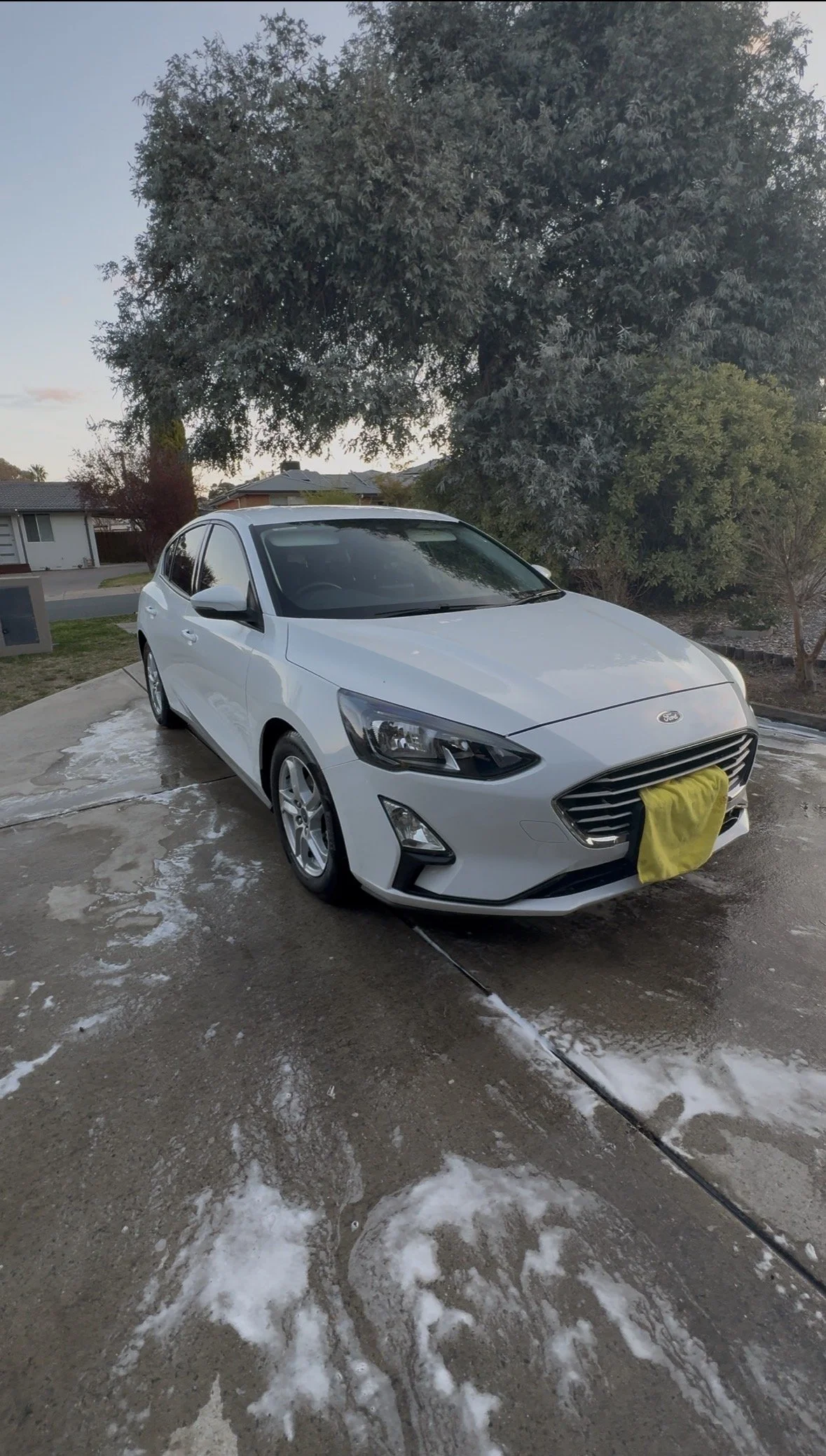 A white sedan car is parked outside on a wet driveway with soap and water, next to a house and large trees, during evening or early morning.