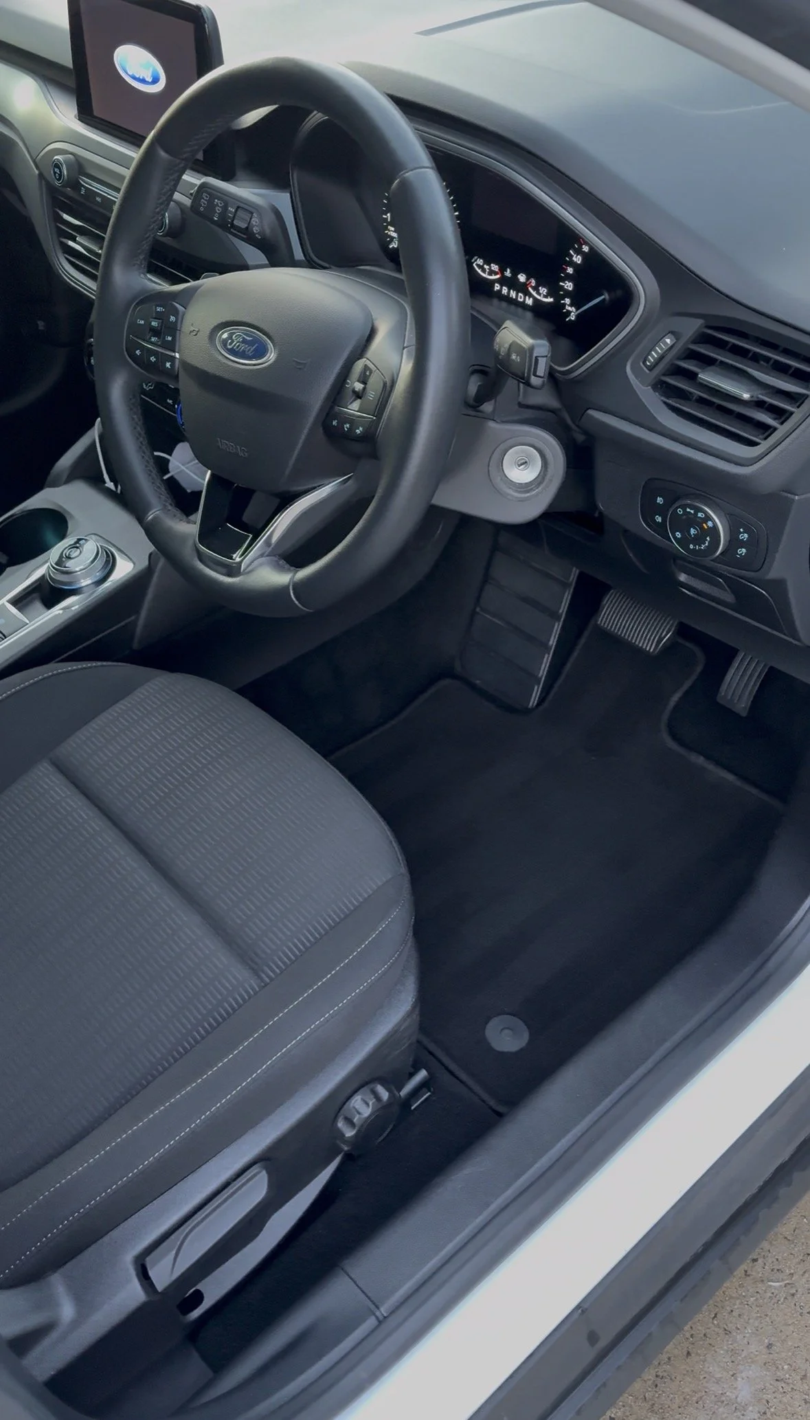 The interior of a Ford vehicle showing the driver's seat, steering wheel with Ford logo, dashboard with speedometer, gear shift, and air vents.