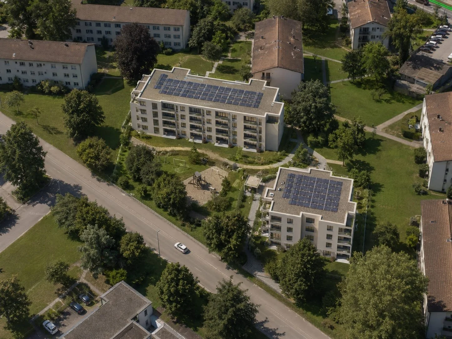 Luftaufnahme eines Wohnviertels mit mehreren Mehrfamilienhäusern, die Solarzellen auf den Dächern haben, umgeben von Bäumen, Gehwegen und Straßen, mit parkenden Autos.
