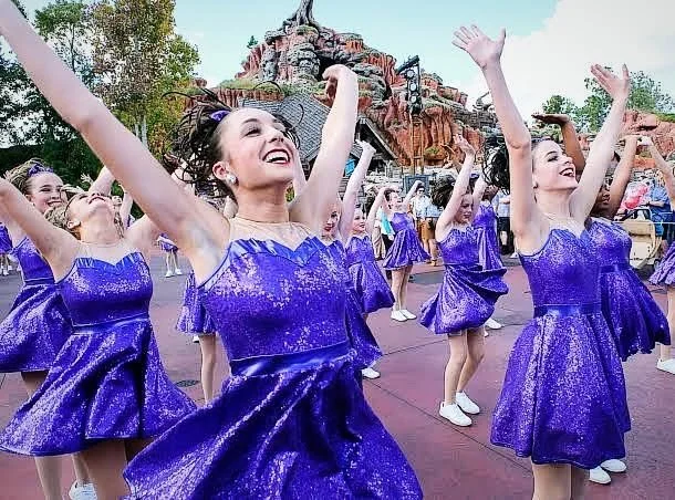 Group of young women in purple dresses dancing outdoors near a castle-themed structure.