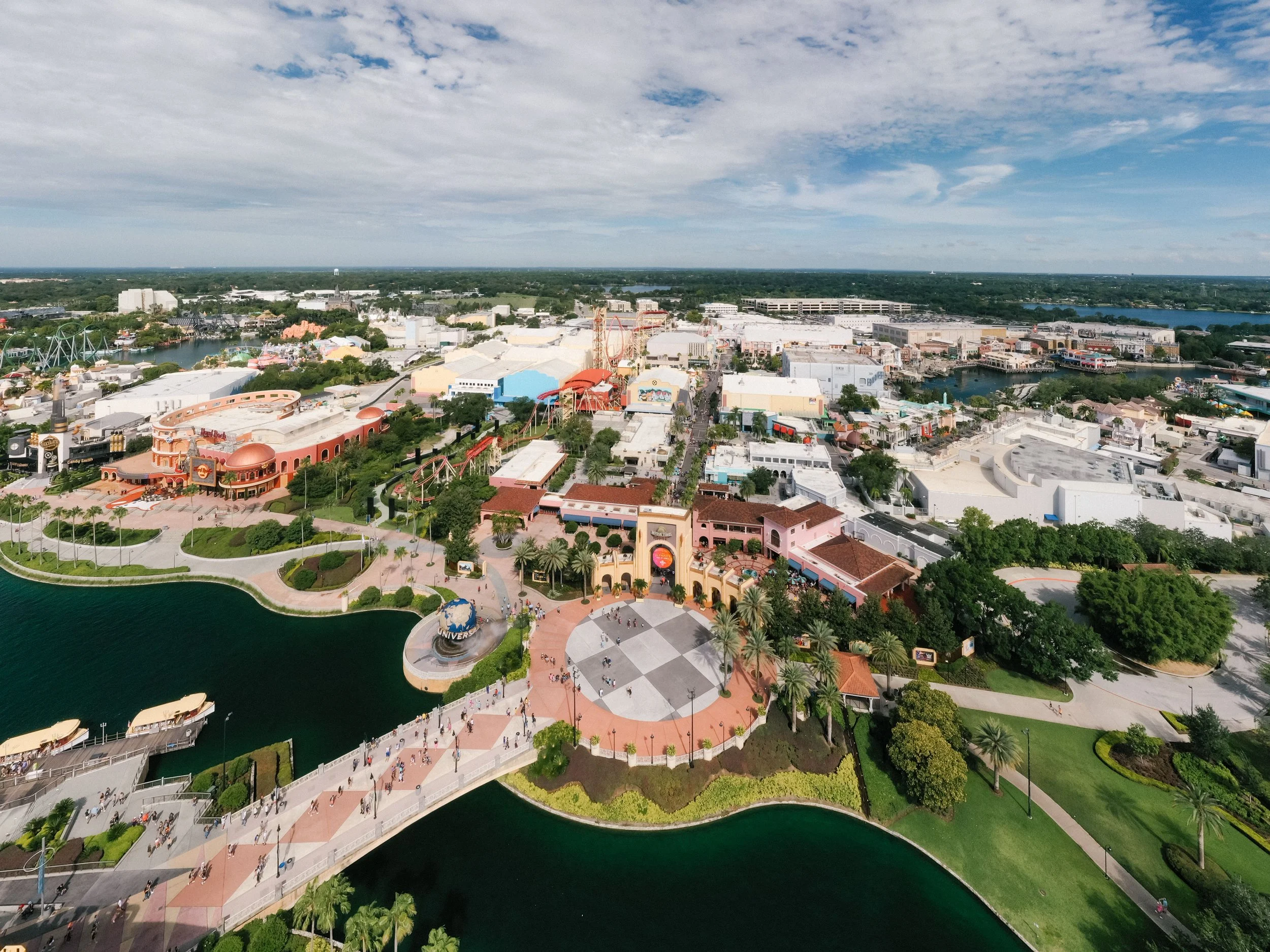 Aerial view of an amusement park with rides, water attractions, a lake, and a cityscape in the distance under partly cloudy skies.