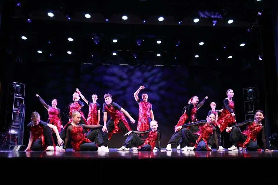 Group of children dressed in red and black dancing on stage with black background and stage lights.