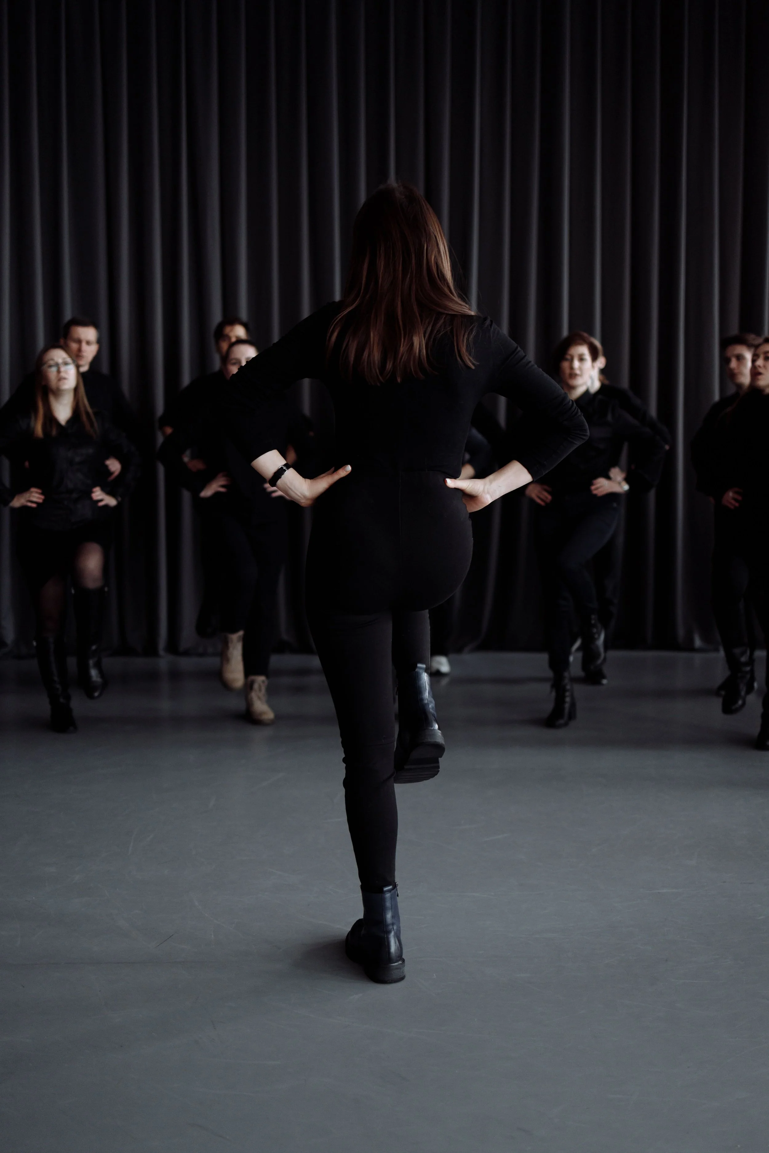 A woman with brown hair dances in front of a group of people on a stage with a dark curtain backdrop.