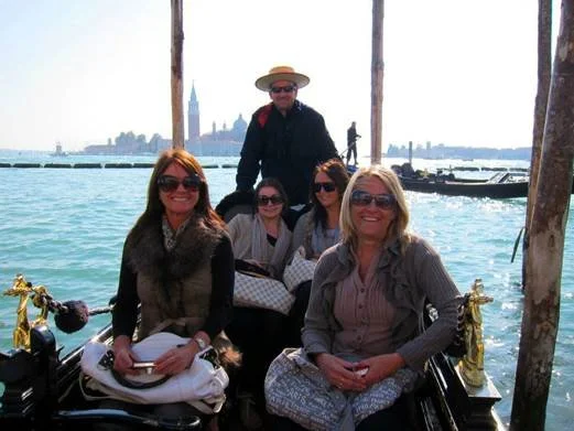 Group of five women and one man on a gondola ride in Venice, Italy, with the city skyline and water in the background.