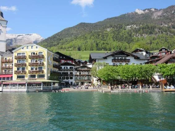 A lakeside view of buildings with mountains in the background, including a hotel named Seeblickhotel, alongside green trees and a clear blue lake.