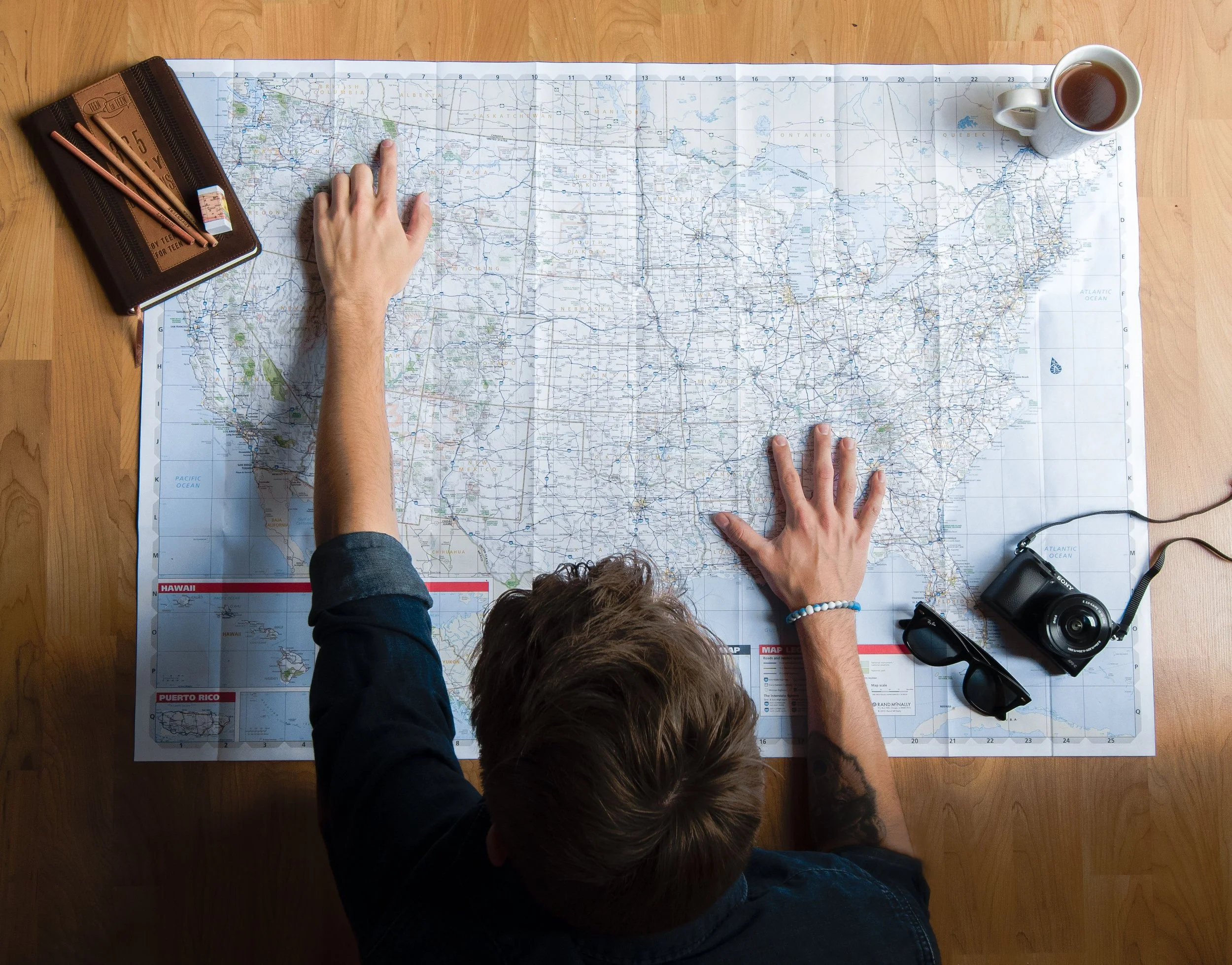 Overhead view of a person with long hair and a dark jacket looking at a large map of the United States laid out on a wooden table. The person's right hand is pointing to North Dakota while their left hand is resting near the map. A coffee mug is on the top right corner, and a camera with a strap is placed to the bottom right. A closed journal or planner with a zipper and some pens is on the left side of the map.