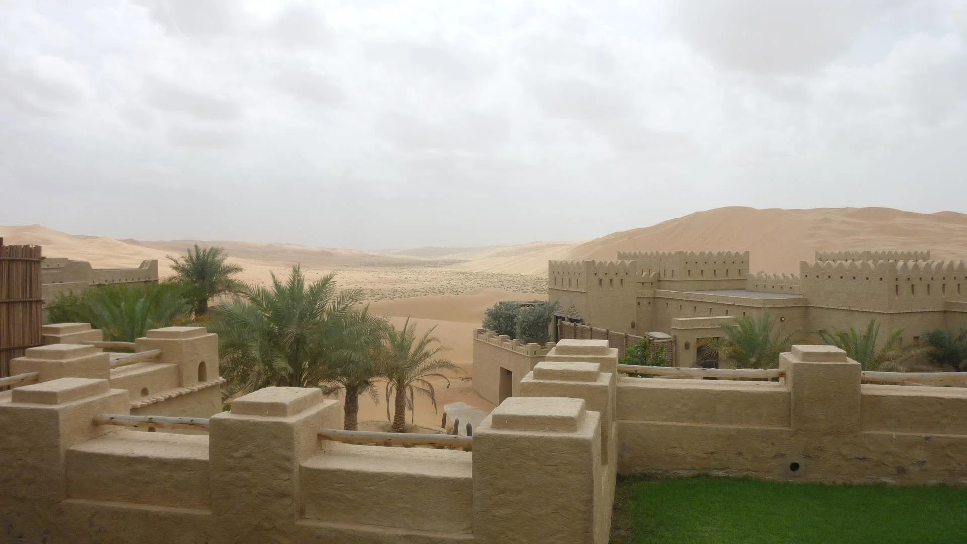 View of a desert landscape with sand dunes, palm trees, and traditional Middle Eastern style buildings with beige walls and crenellated rooftops.