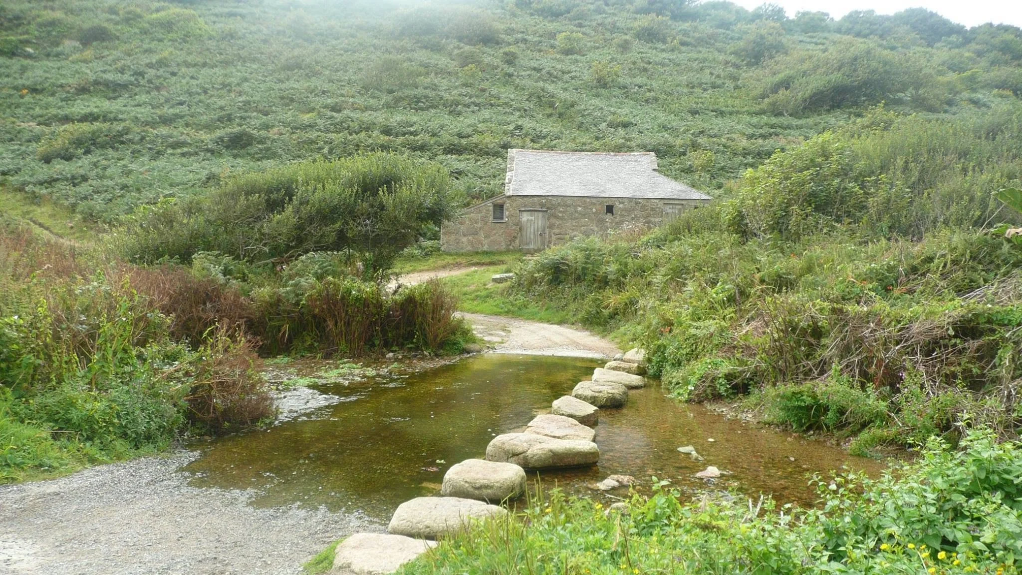 A peaceful scene of a small stone house with a pitched roof in a lush green hillside, next to a narrow stream with stepping stones. There is a gravel path leading to the house, surrounded by dense vegetation and shrubs.