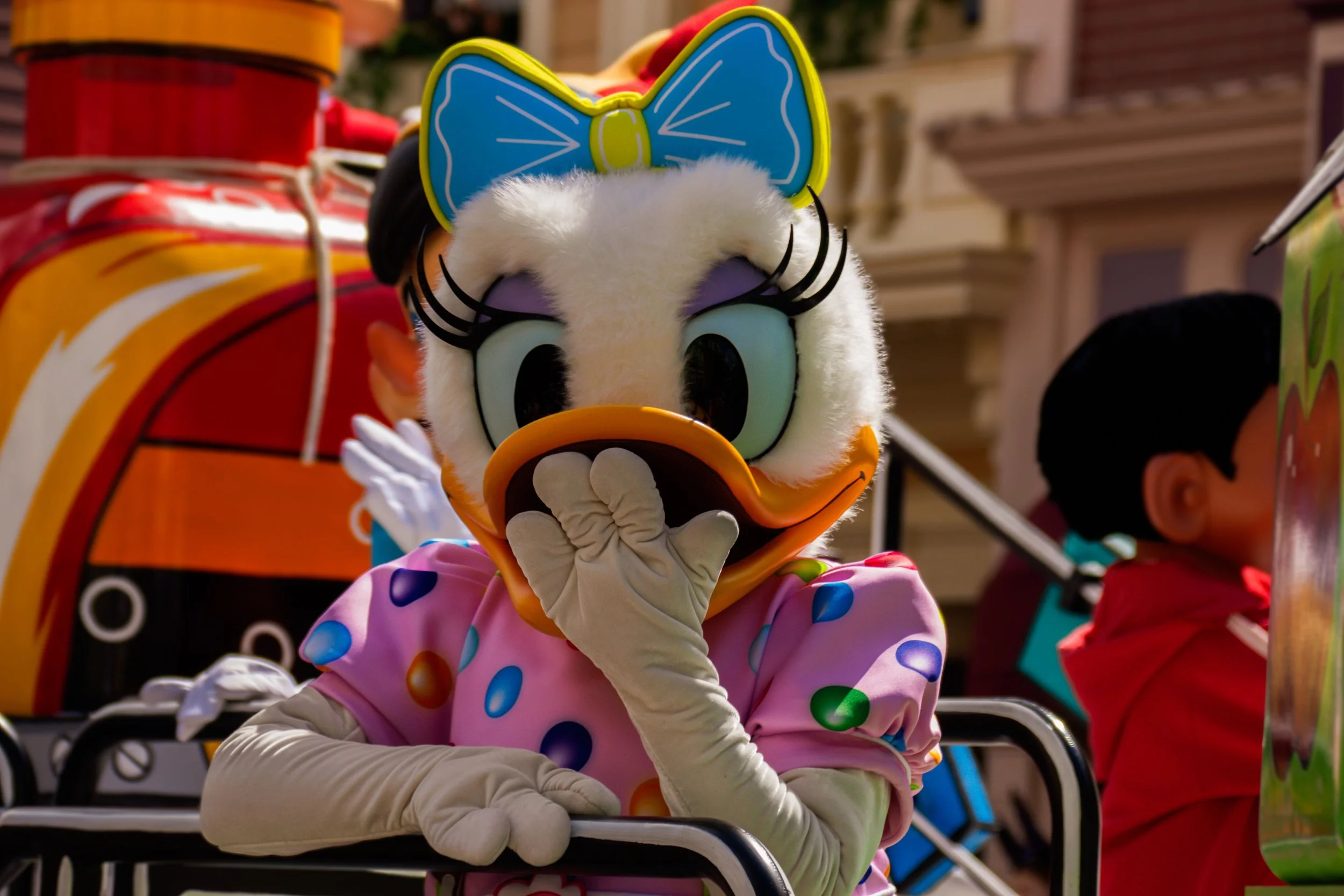Costumed character of Daisy Duck with a blue bow on her head, wearing pink polka-dot dress, sitting on a parade float with hand on chin.