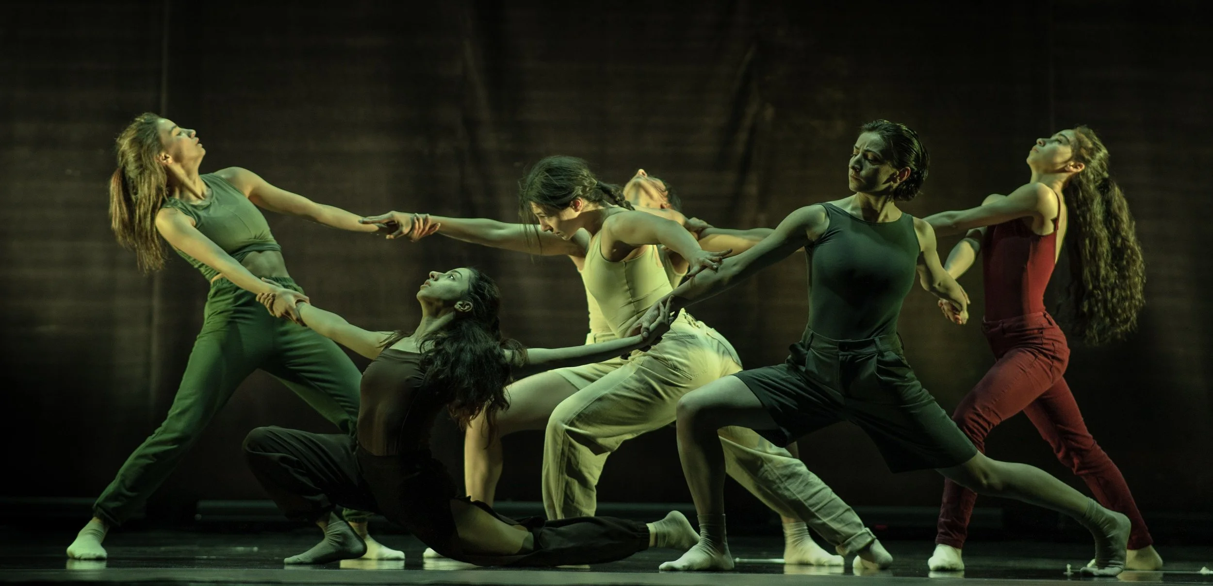A group of six dancers performing on stage, holding hands, with some in kneeling or crouching positions, wearing casual attire and expressive in their movements.