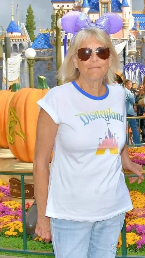 A woman with blonde hair wearing Mickey Mouse ears and sunglasses, standing in front of Disneyland castle, with large pumpkins and fall decorations around.