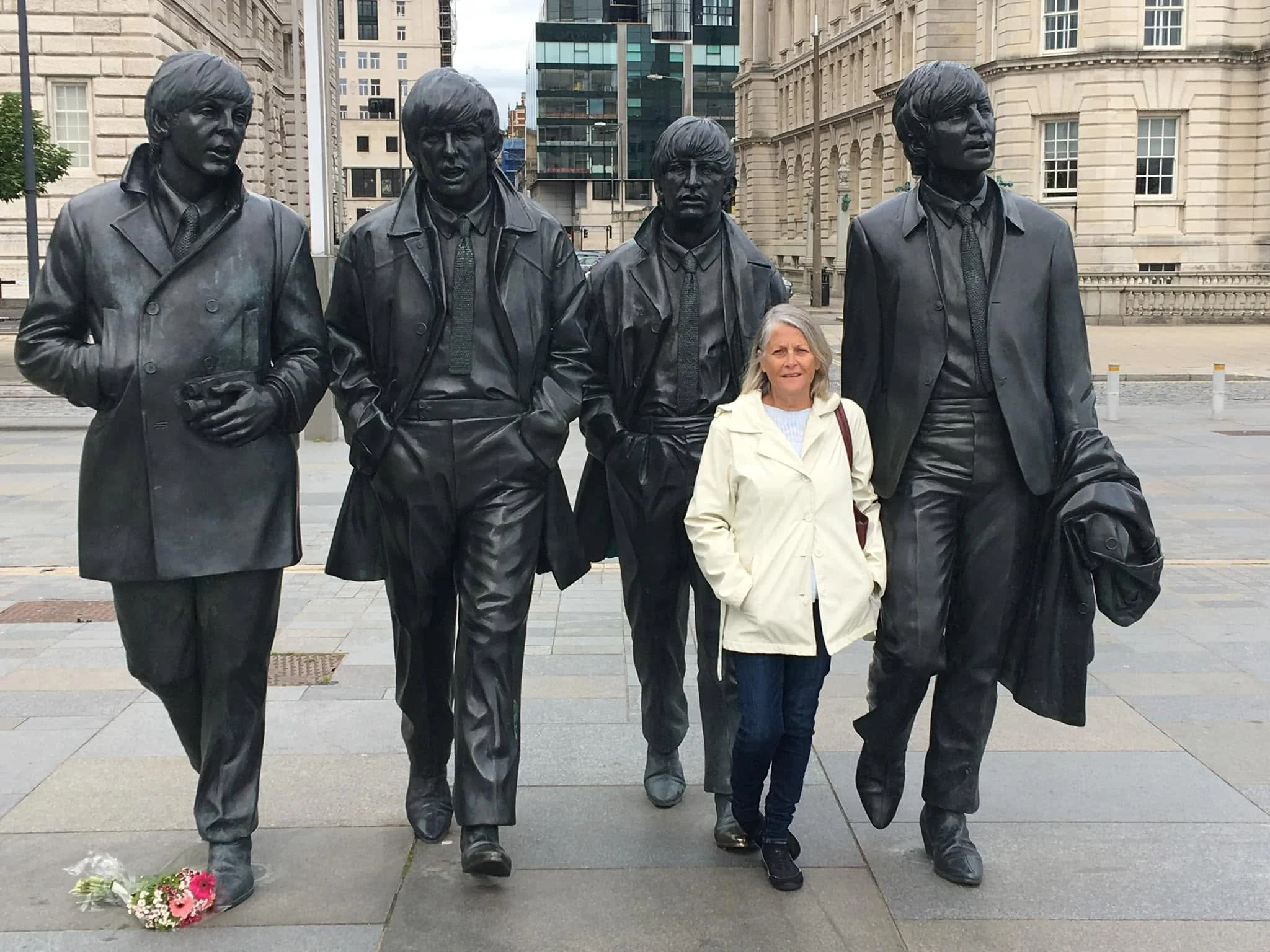 A woman standing among four statues of The Beatles, which are life-sized and made of bronze. The statues depict the band members in mid-performance, dressed in 1960s-style clothing. The woman appears to be in a city square, with modern and classical 