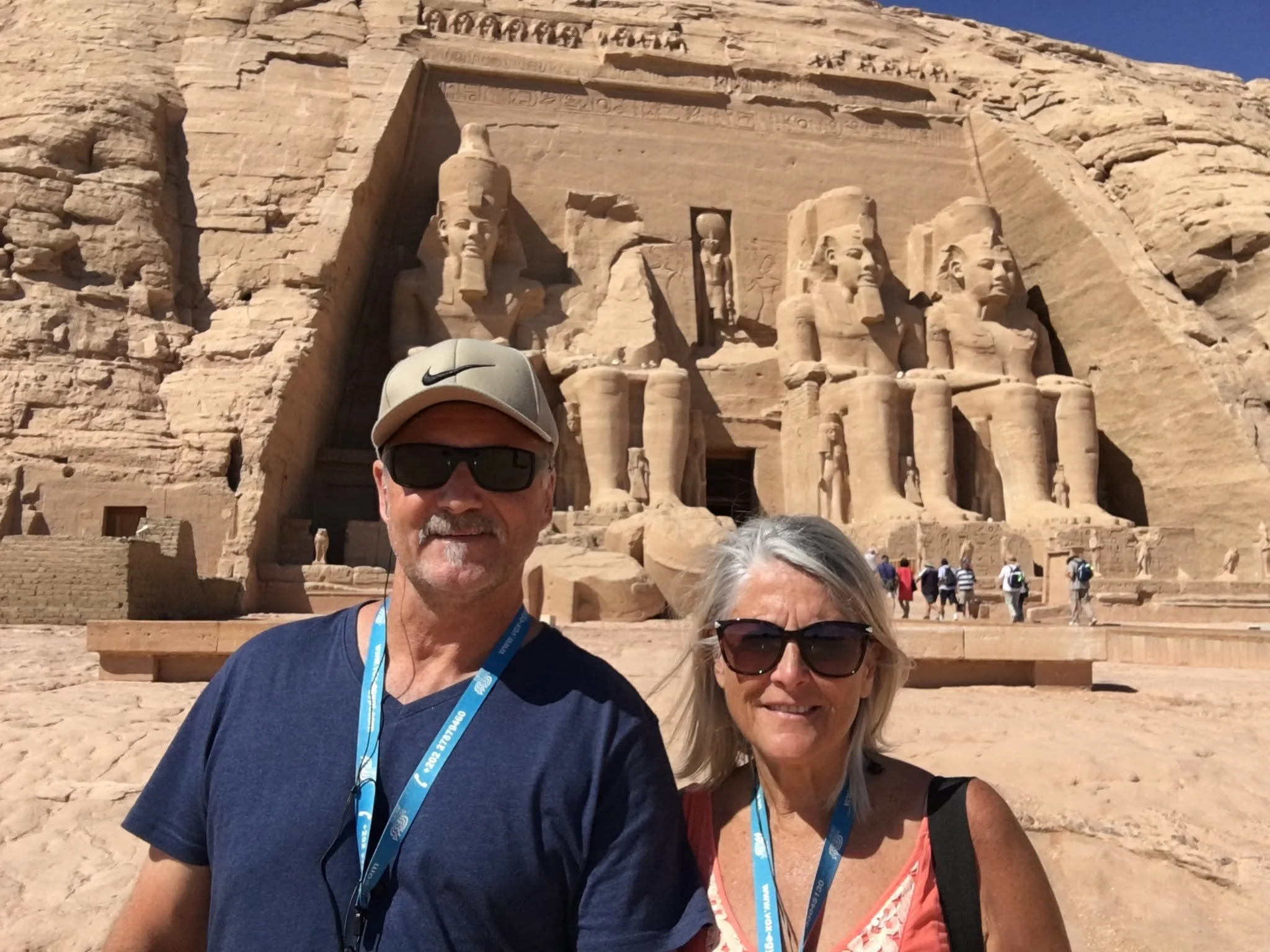 Two smiling tourists, a man and a woman, pose in front of the ancient Egyptian Abu Simbel temples carved out of the rock cliff in Egypt. The temples are decorated with large statues of Egyptian gods and pharaohs.