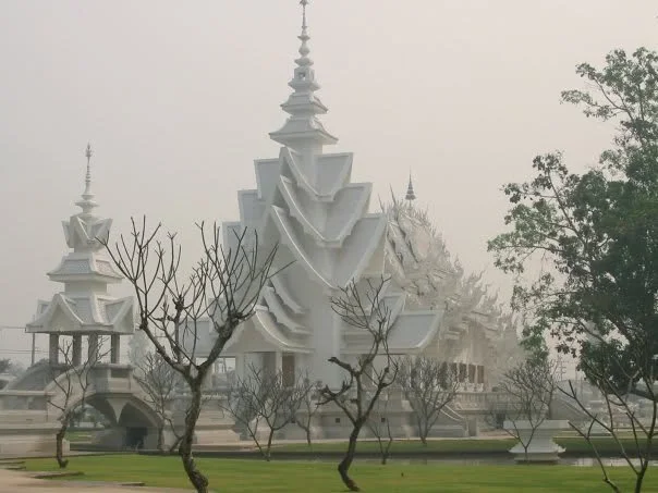 A large, white, ornate building with traditional Asian architecture, featuring multiple spires and curved roofs, set against a foggy sky with leafless trees in the foreground.