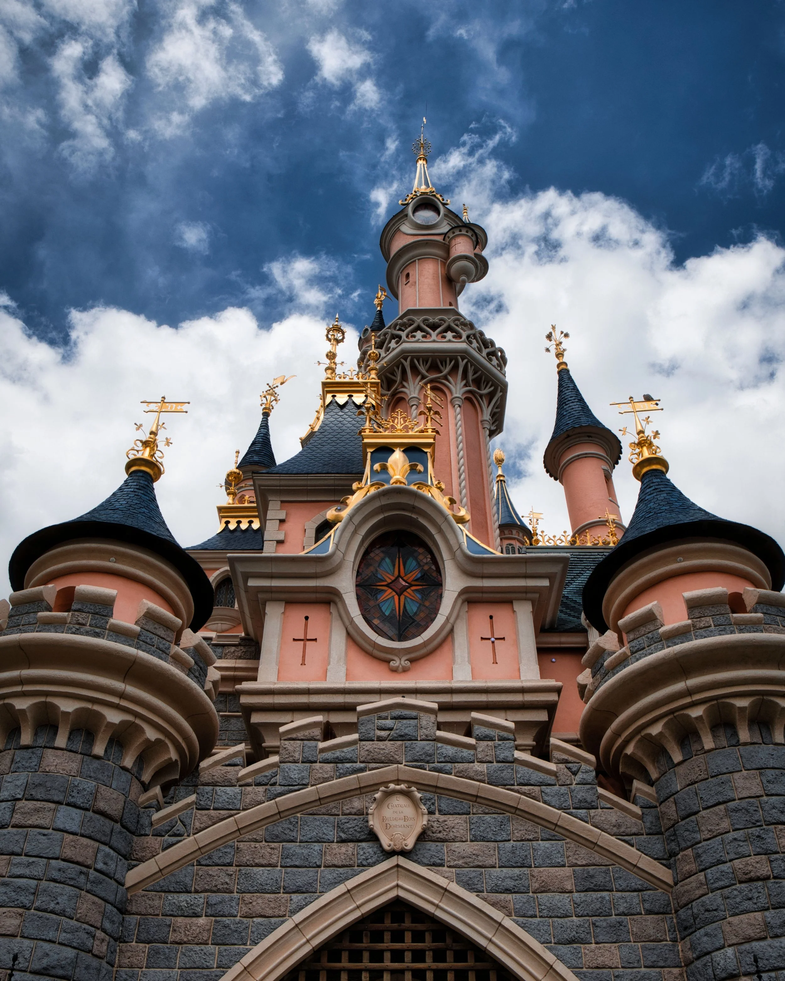 A pink castle with turrets topped with blue and gold roofs, decorative gold accents, and a stained glass window, set against a partly cloudy sky.