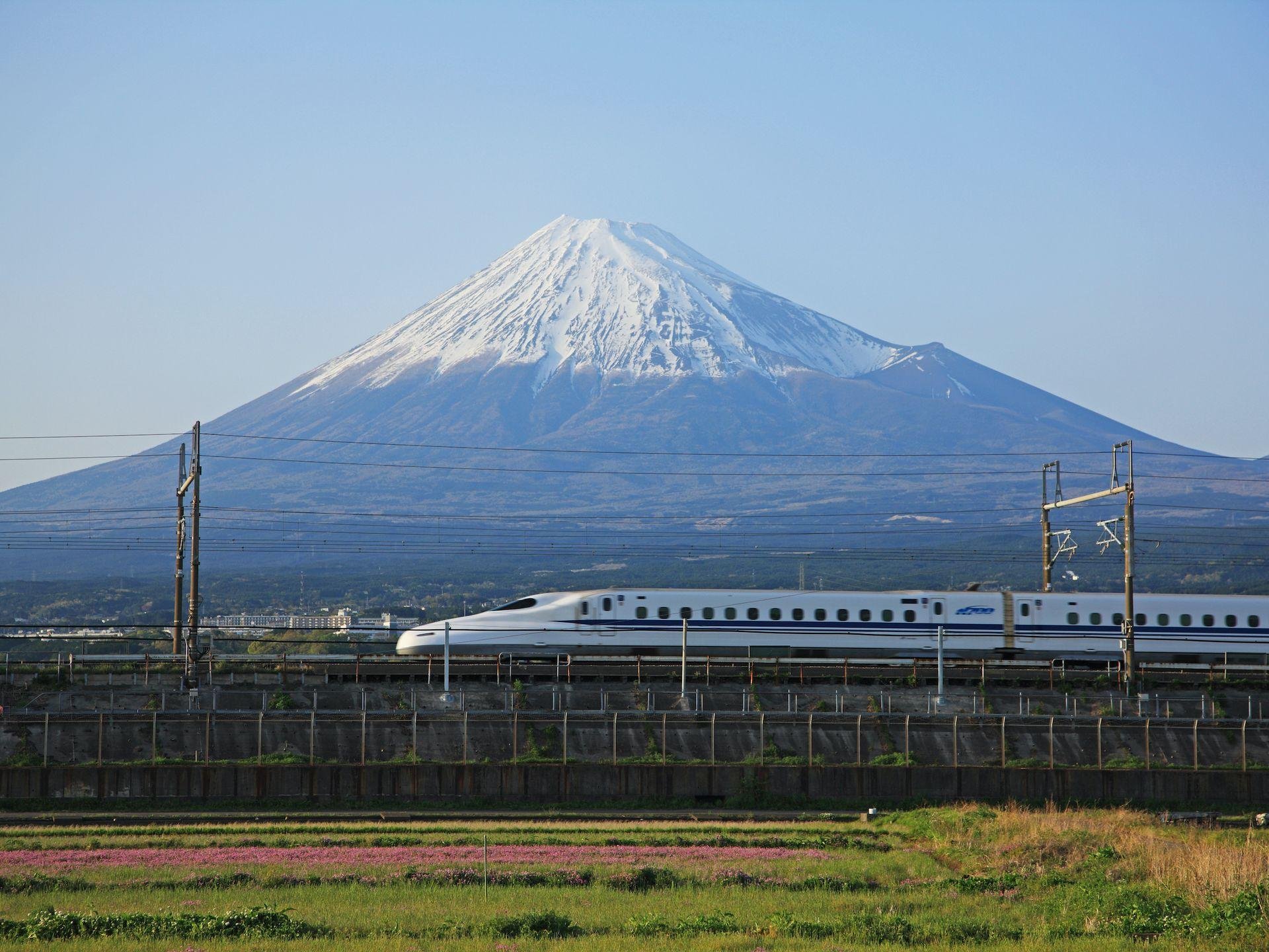 Snow-capped Mount Fuji seen from a distance with a bullet train passing in the foreground and power lines across the scene.