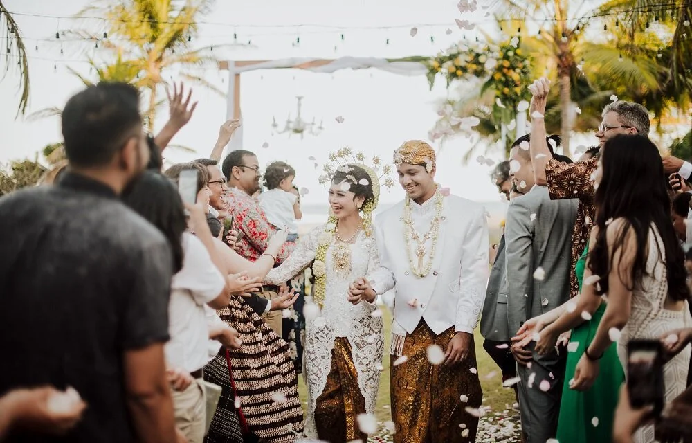 A traditional outdoor wedding ceremony with the bride and groom in traditional attire, surrounded by friends and family throwing flower petals, with palm trees and string lights in the background.