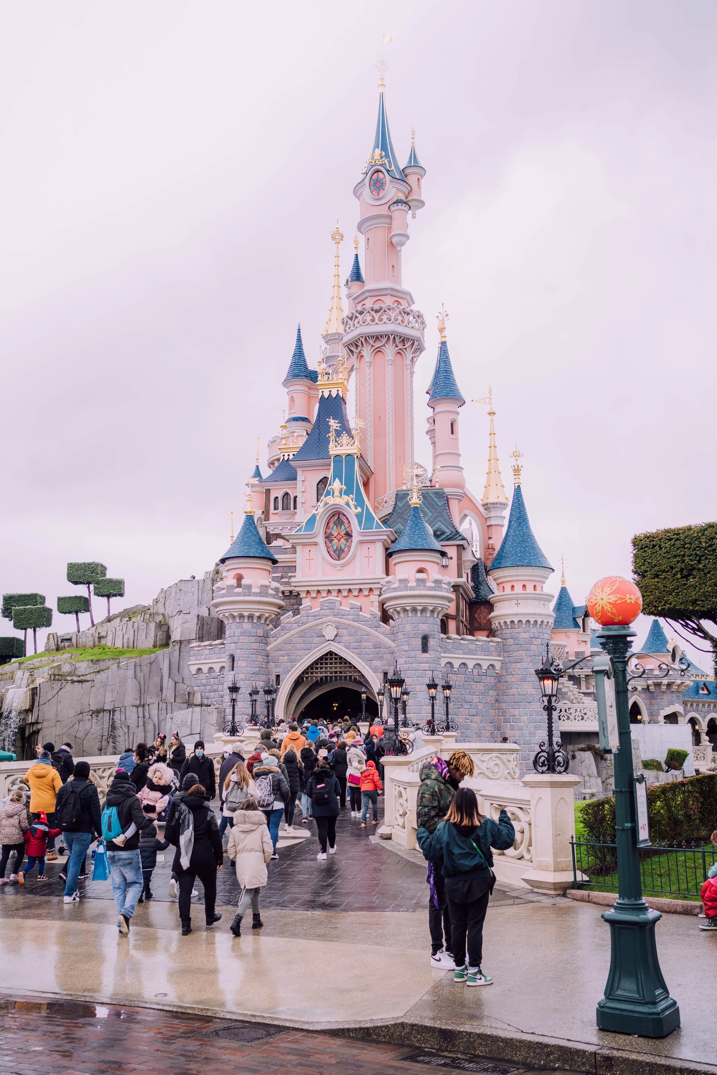 People walking towards Sleeping Beauty Castle at Disneyland, with cloudy sky and wet pavement.