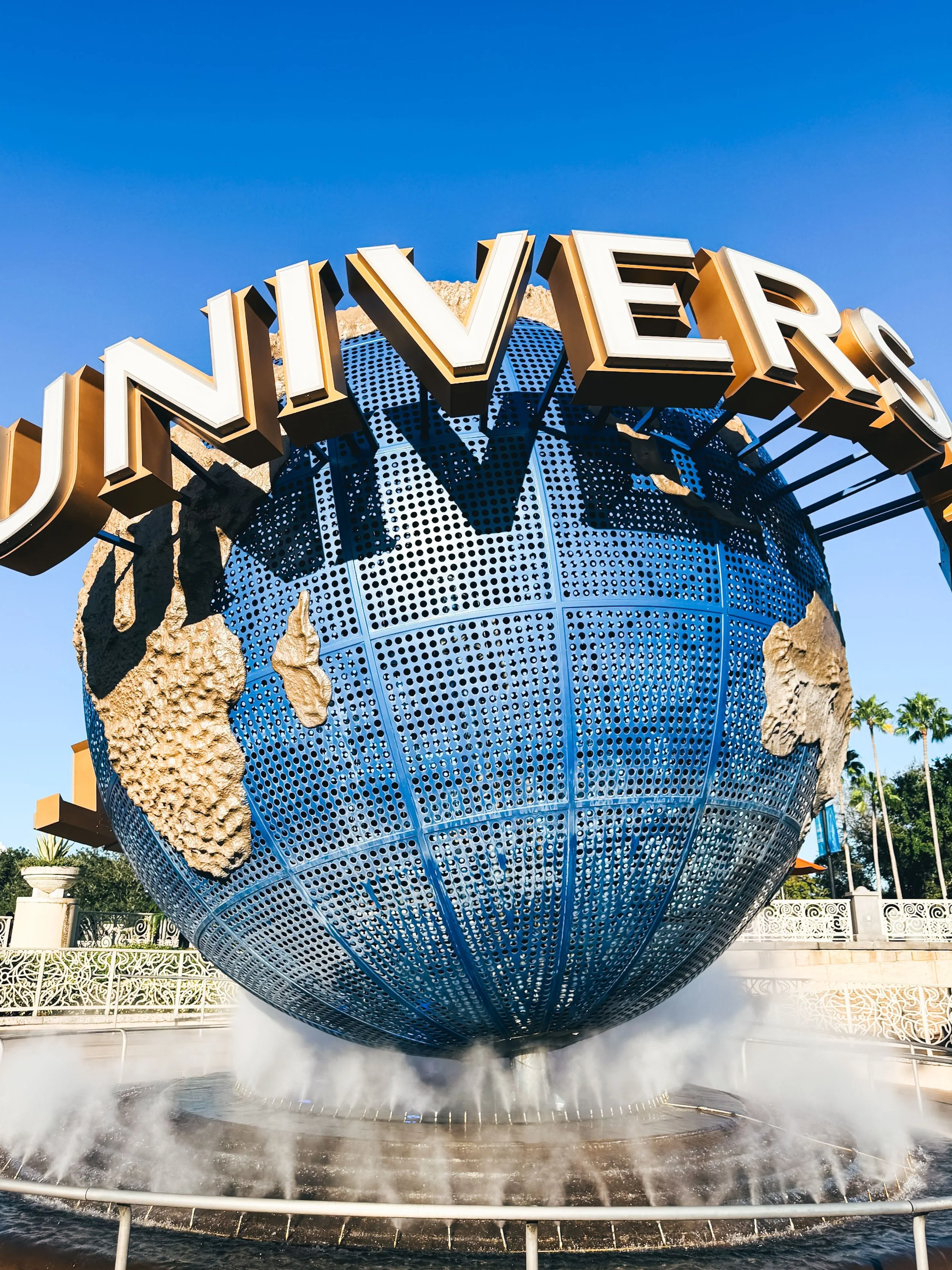 The Universal Studios globe fountain with a large globe featuring cartoonish landmasses and the word 'UNIVERSAL' in large white and gold letters on top, set against a blue sky with some trees in the background.