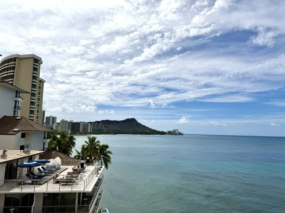 A tropical ocean view with a city skyline and a mountain in the background, featuring a building with an outdoor deck, lounge chairs, and palm trees.