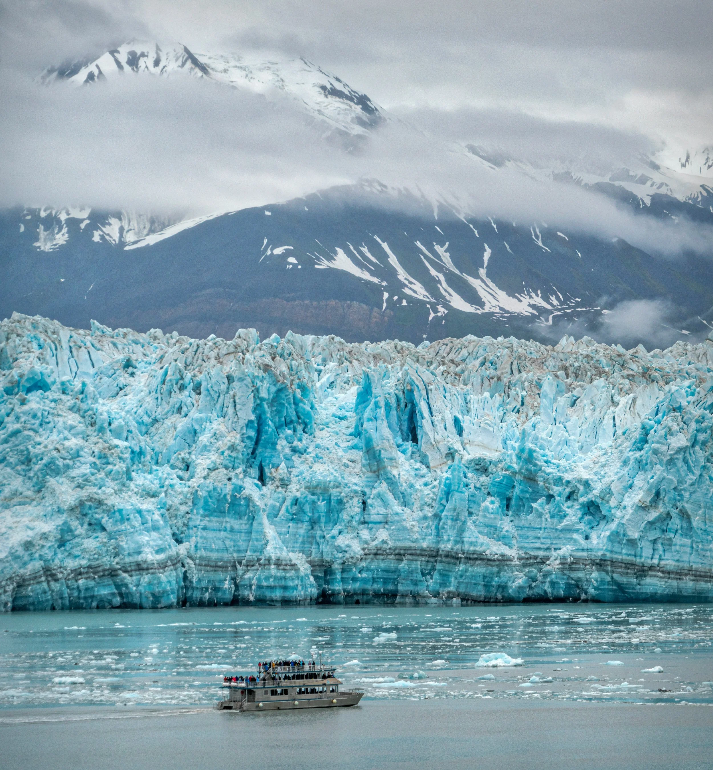 Tourist boat with people observing a glacier and snow-capped mountain through fog and clouds.