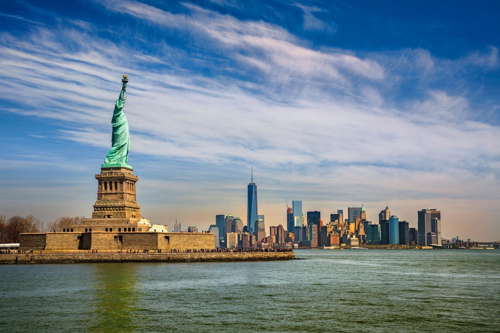 The Statue of Liberty in the foreground with the New York City skyline, including One World Trade Center, in the background under a partly cloudy blue sky.