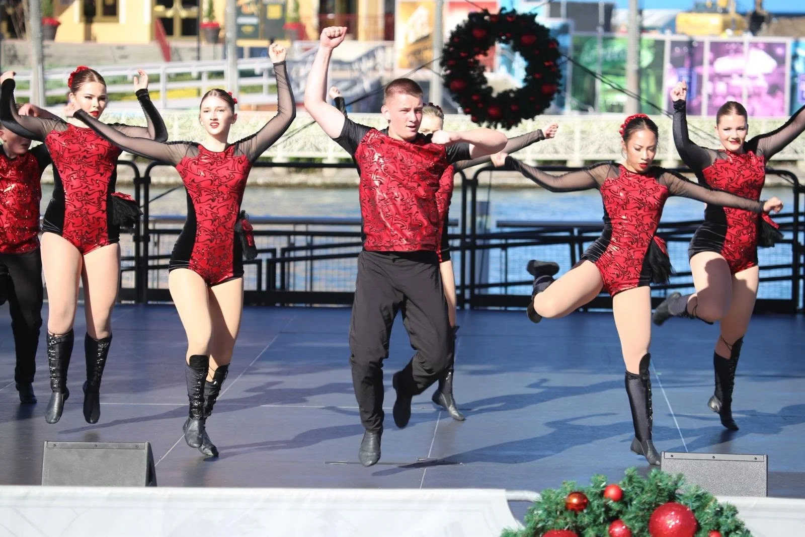 A group of dancers performing on an outdoor stage, dressed in red and black costumes, with Christmas decorations in the foreground and a festive wreath hanging in the background.