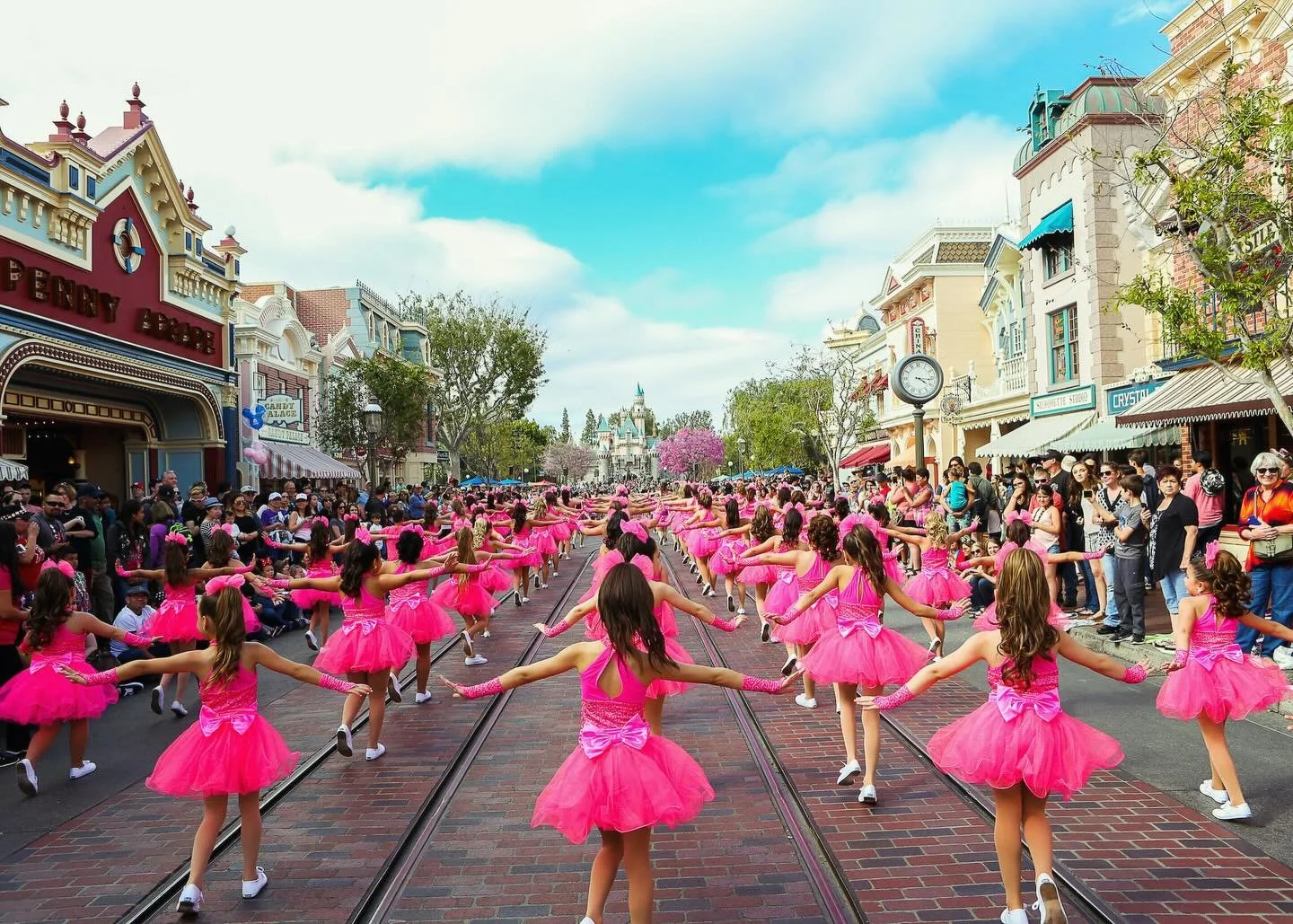 A parade of young girls in pink dresses dancing on streetcar tracks in a theme park, with audience watching on both sides, and colorful buildings and a castle in the background.