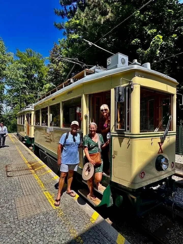 Three people stand on the steps of a vintage yellow streetcar with a woman inside. The streetcar is on a shadowed track, surrounded by trees, with a bright blue sky overhead.