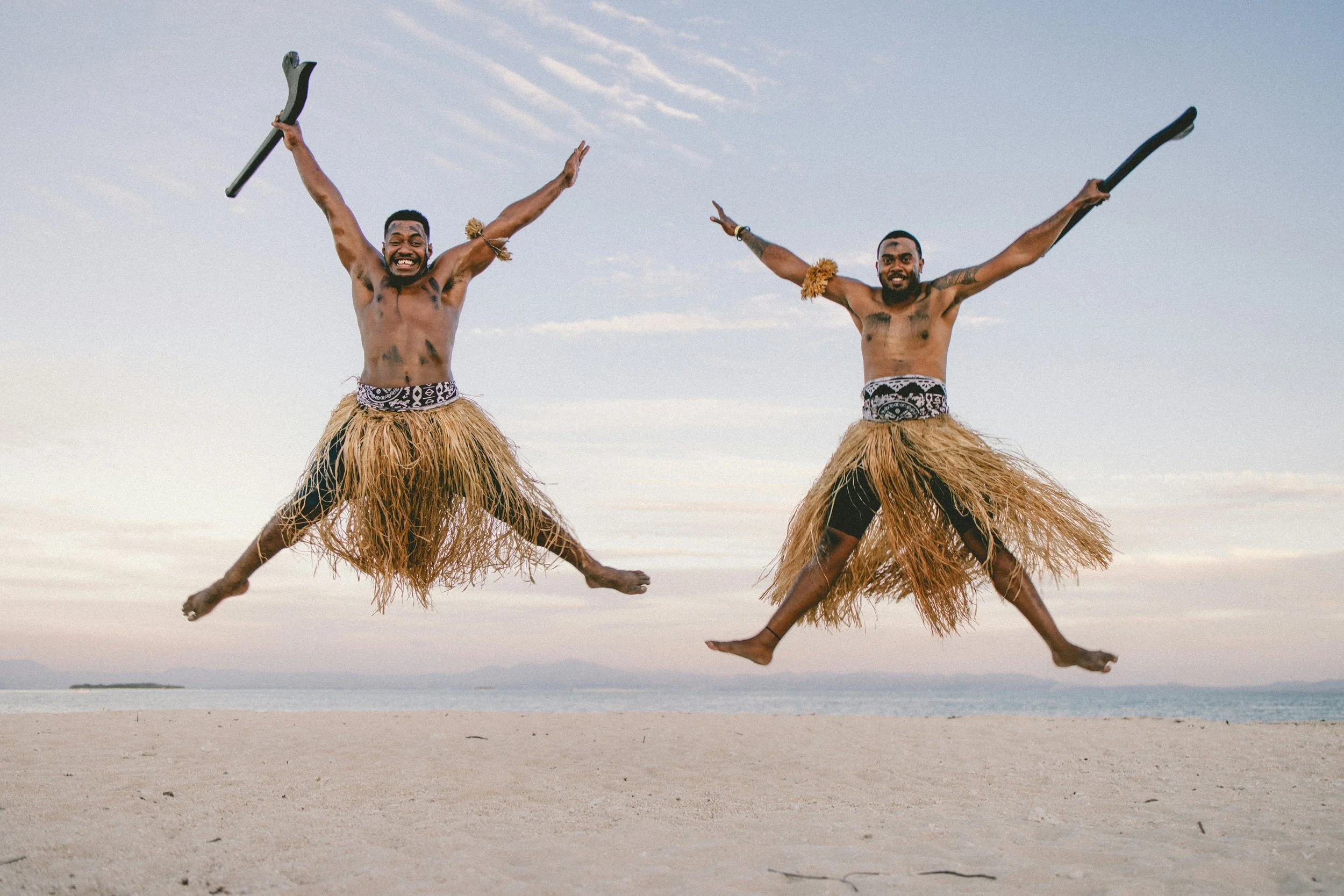 Two men wearing grass skirts jumping in the air on a beach, holding sticks, with arms outstretched and smiling, during daytime with a cloudy sky.