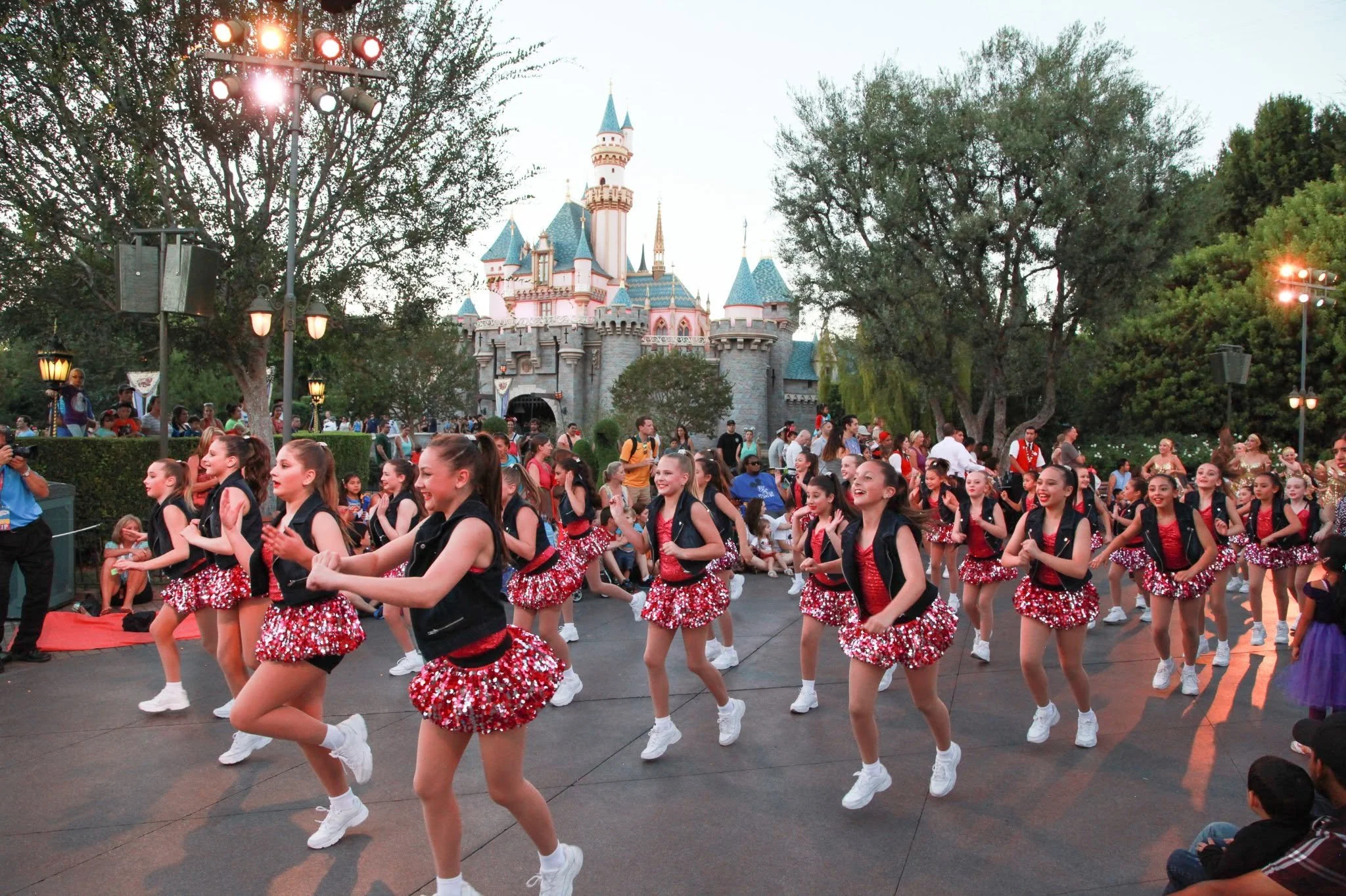 A parade of young dancers dressed in black and red costumes with sparkly skirts and white shoes marching in front of Sleeping Beauty Castle at Disneyland.