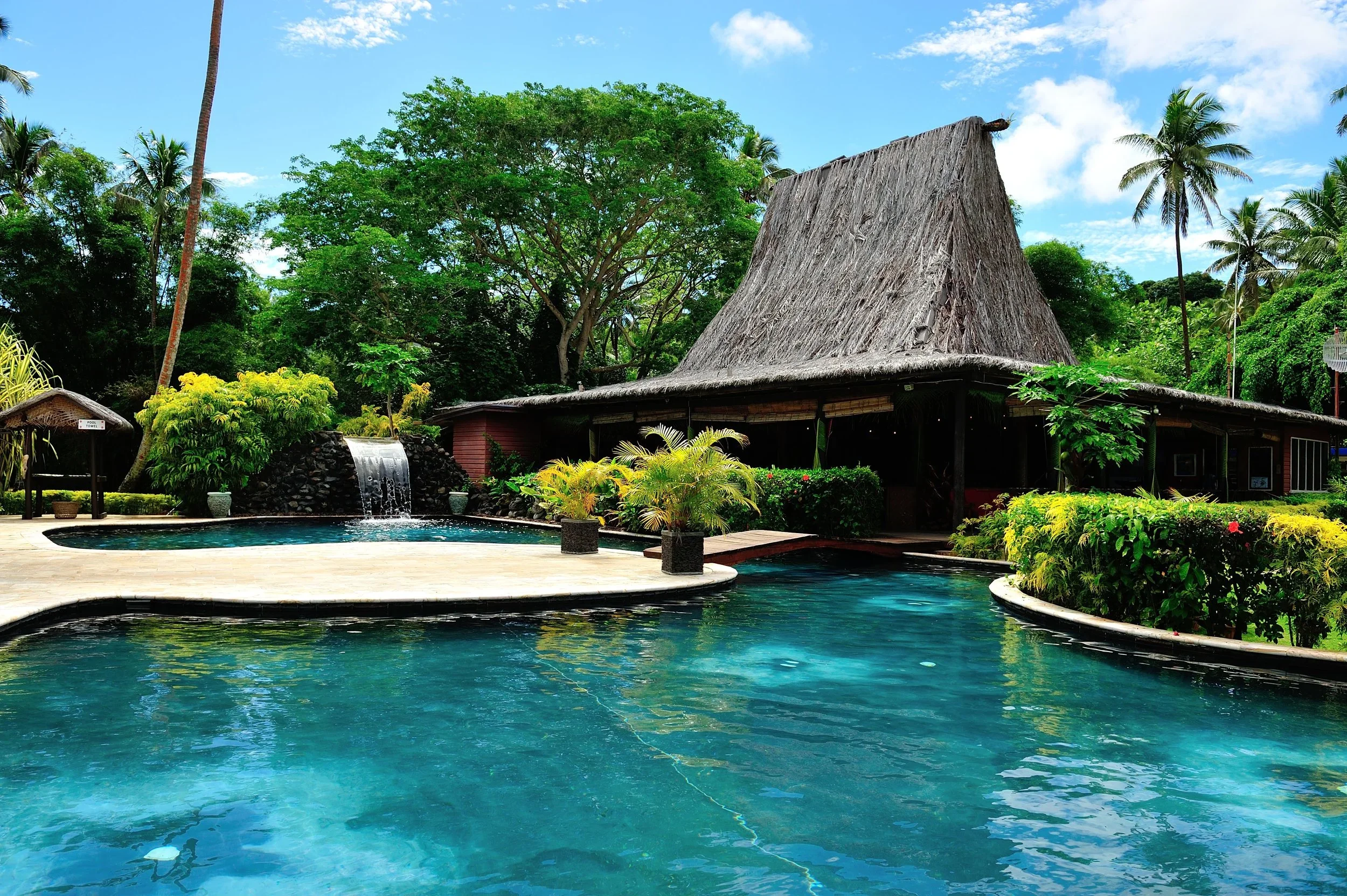 A tropical scene with a thatched-roof hut surrounded by lush green trees, a swimming pool, and a waterfall feature under a blue sky with some clouds.