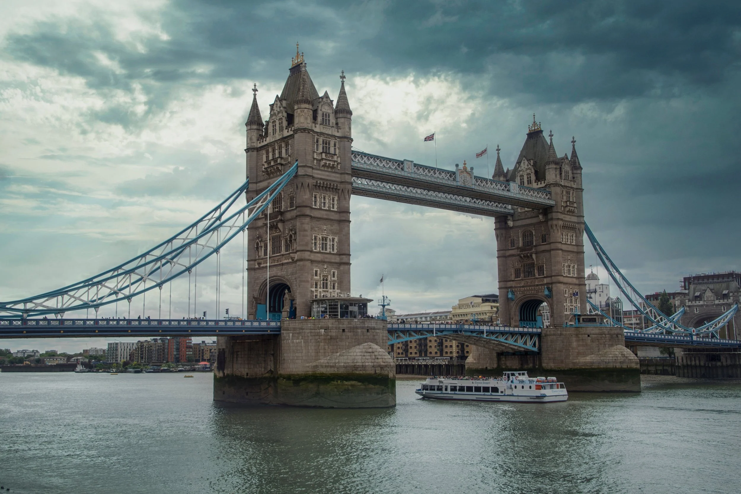 The Tower Bridge in London, England, with a boat sailing on the River Thames beneath it, and cloudy sky above.