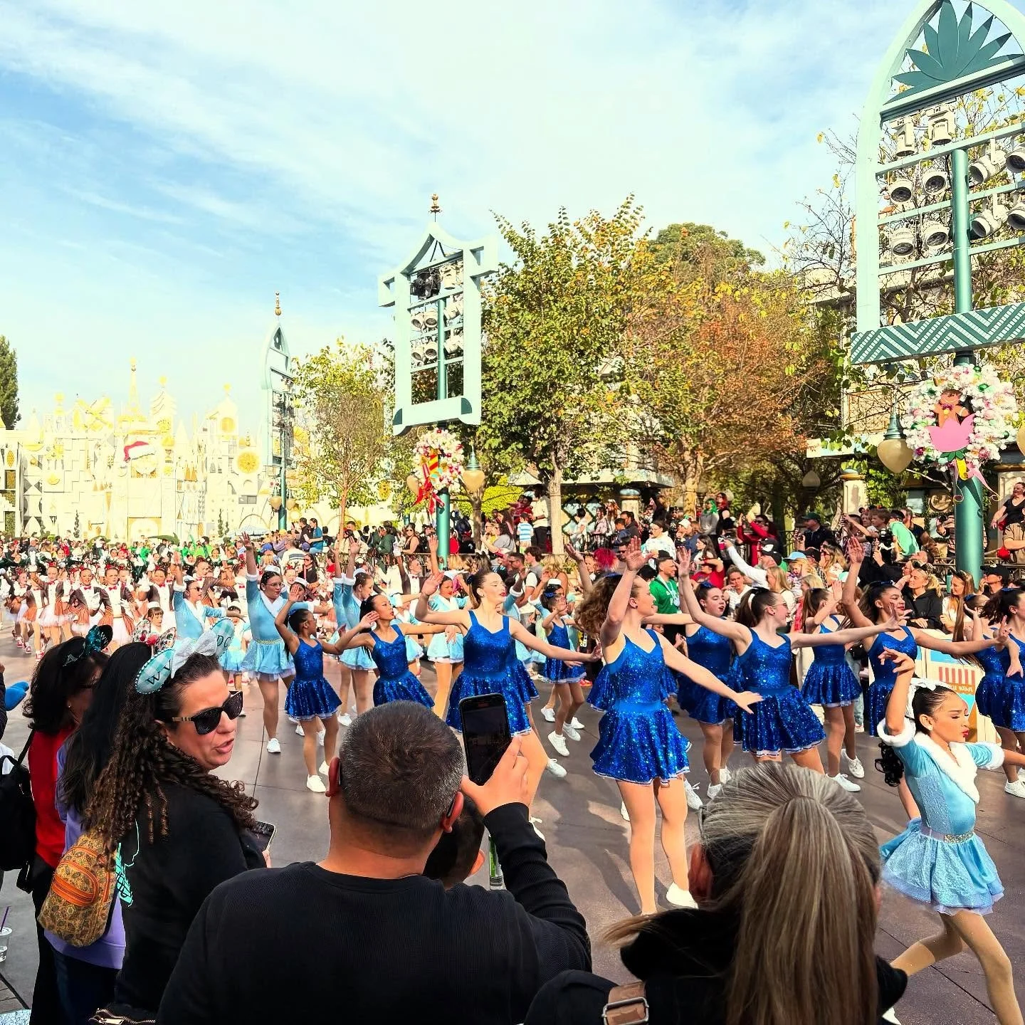Parade of dancers performing in blue costumes at a theme park with onlookers watching.