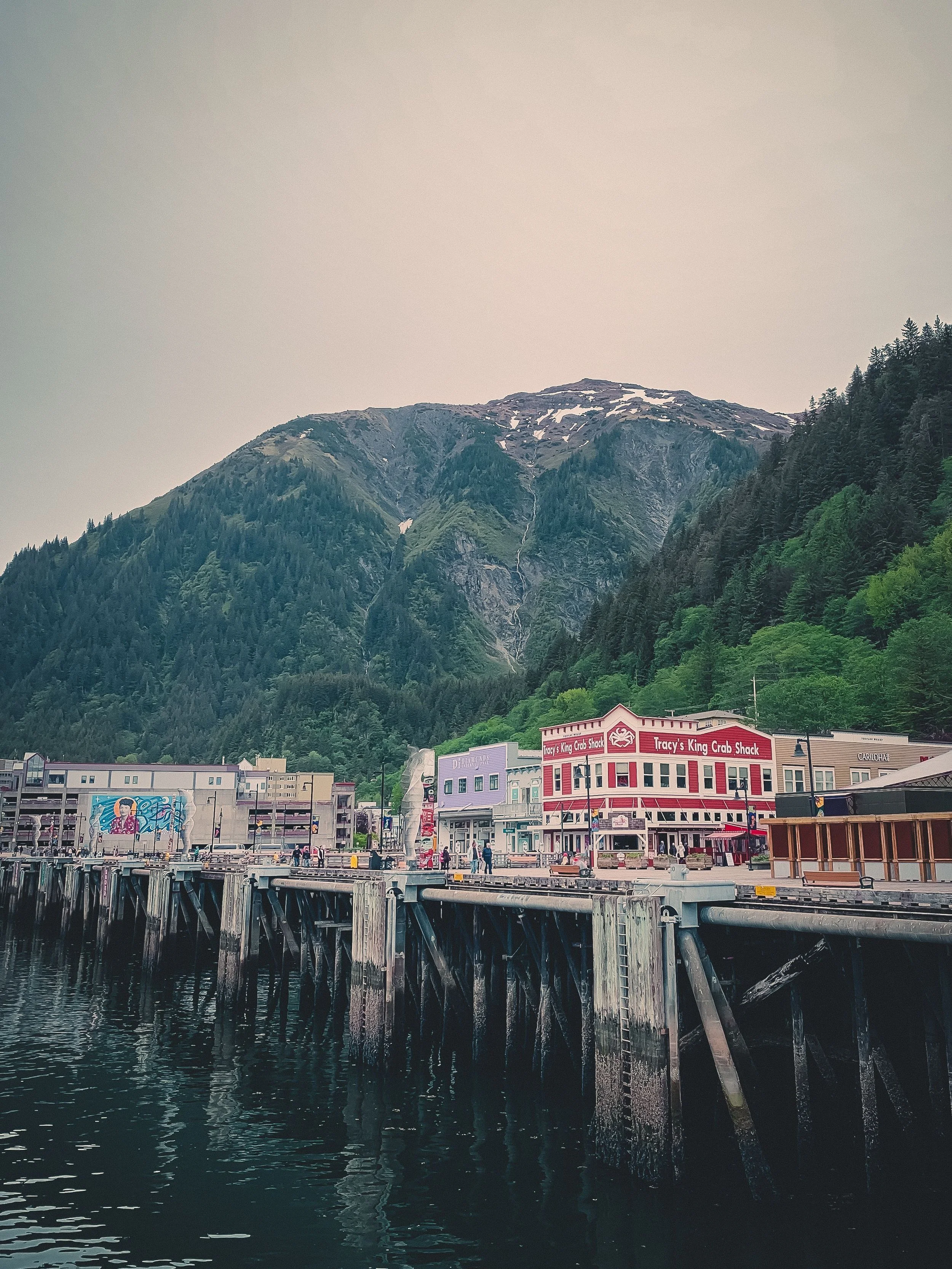 A waterfront town with commercial buildings, including Tracy's King Crab Shack, in front of lush green mountains with patches of snow at the peaks. The harbor has wooden pilings and water reflecting the scene.