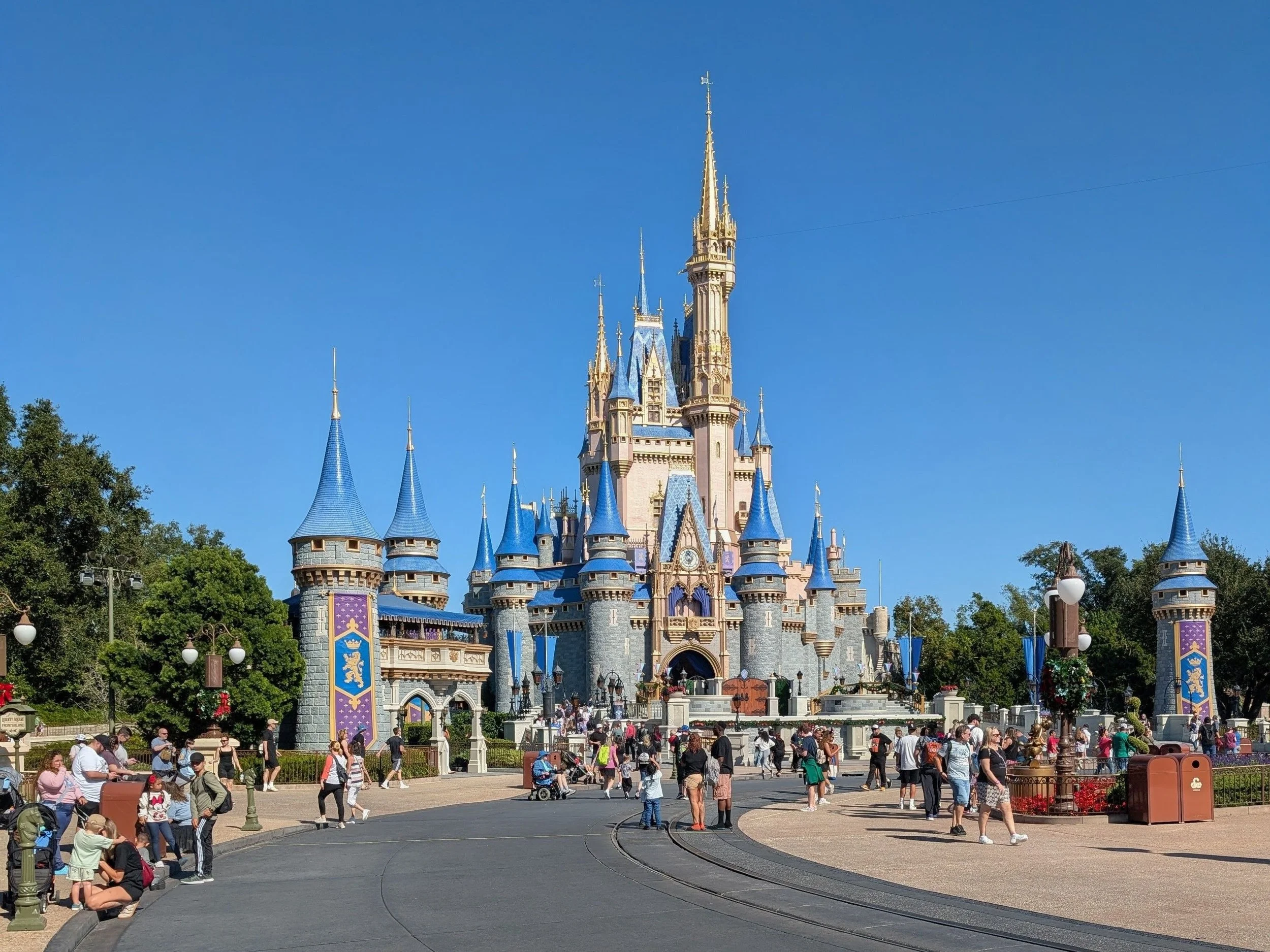 Disney castle with blue rooftops and a crowd of visitors walking and sitting in front on a sunny day.
